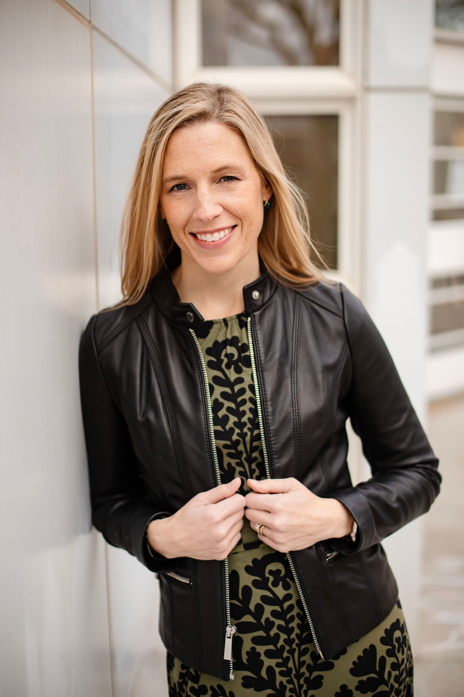 Professional portrait in black leather jacket and patterned dress against white exterior wall.