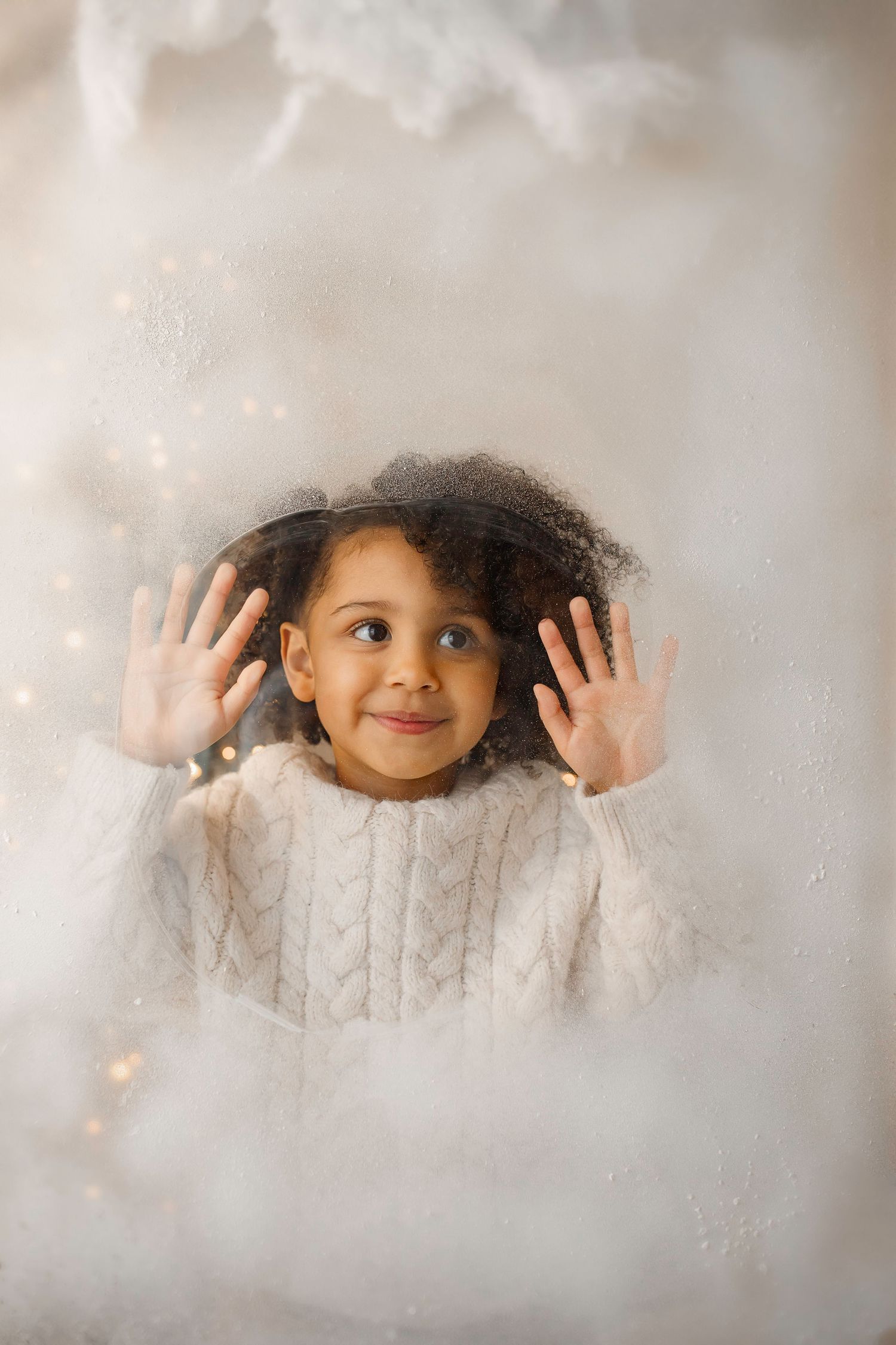 Child in white sweater playfully pressing hands against frosted window with bokeh lights.