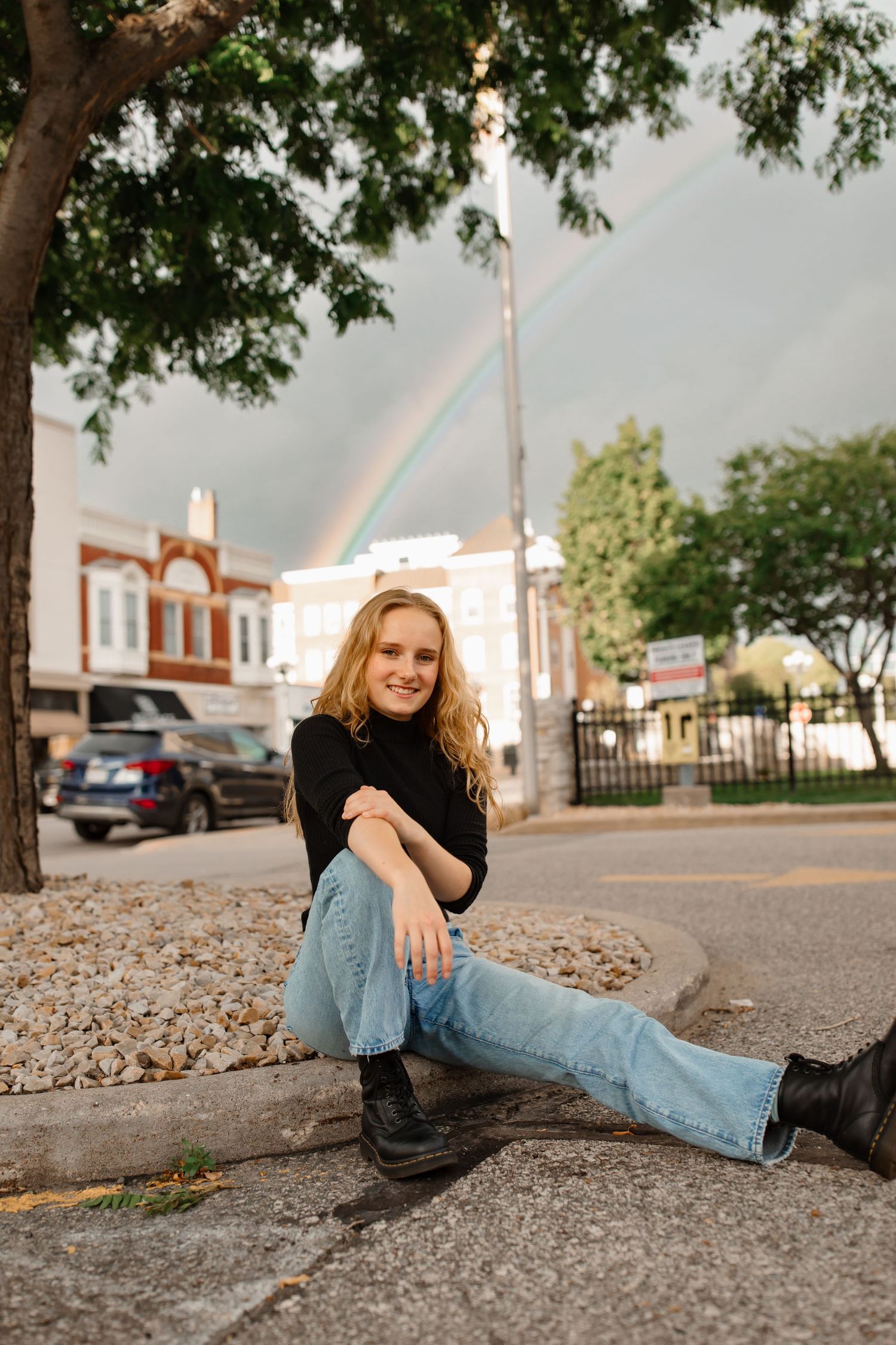 A casual outdoor portrait with blue jeans and black sweater against a rainbow in the sky.