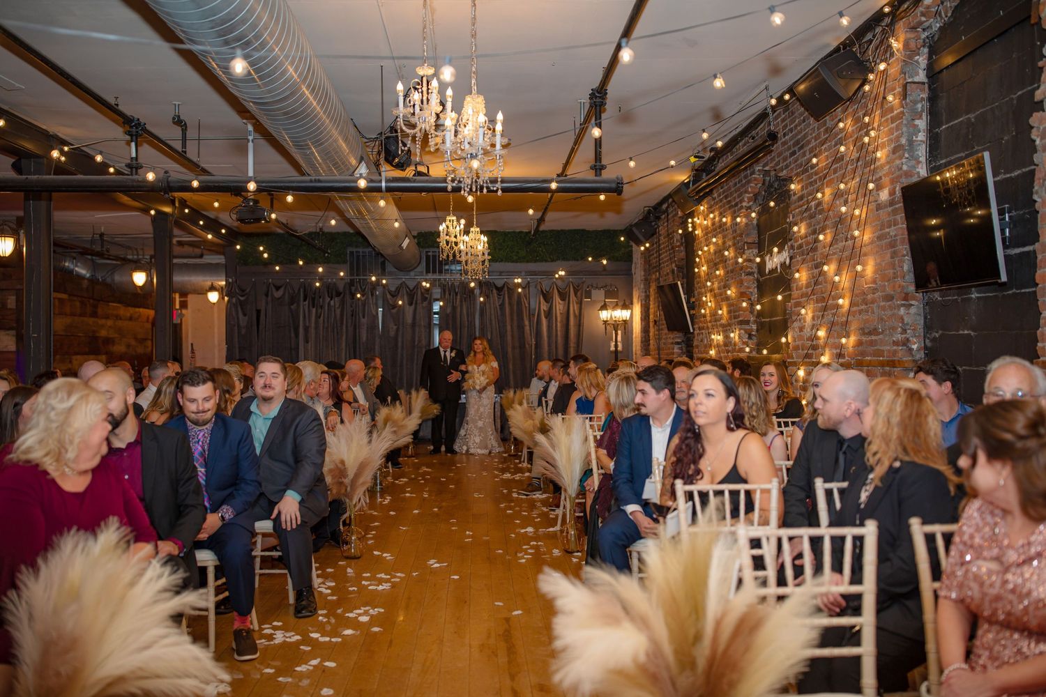 Wedding reception venue decorated with chandeliers and string lights as guests fill white chairs.