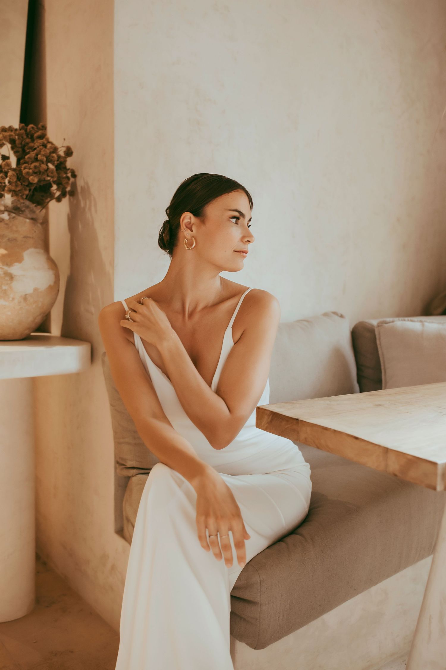 Woman in white dress seated elegantly at dining table in warm minimalist setting.