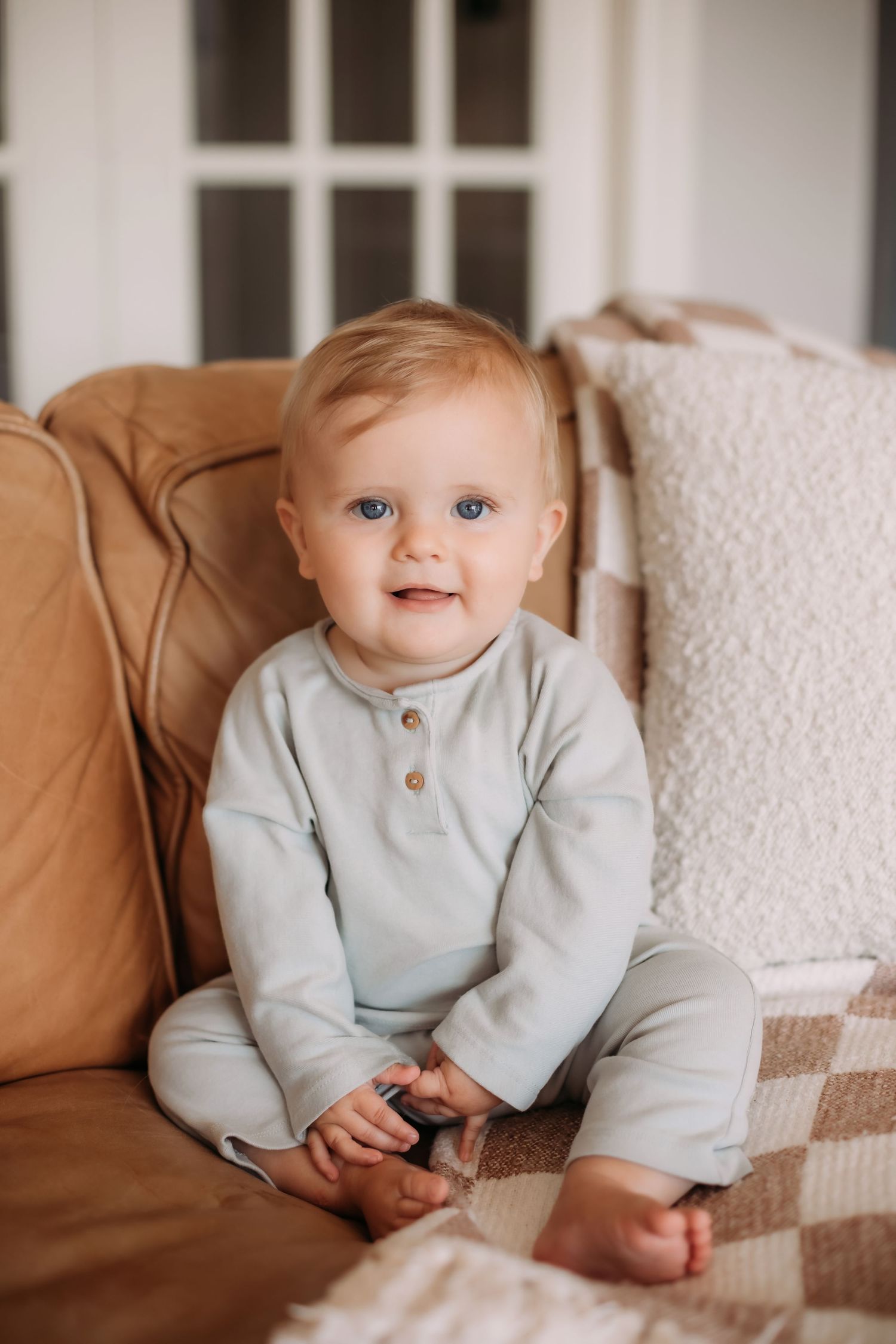 A baby in light blue outfit sits on a tan couch with a sweet smile.