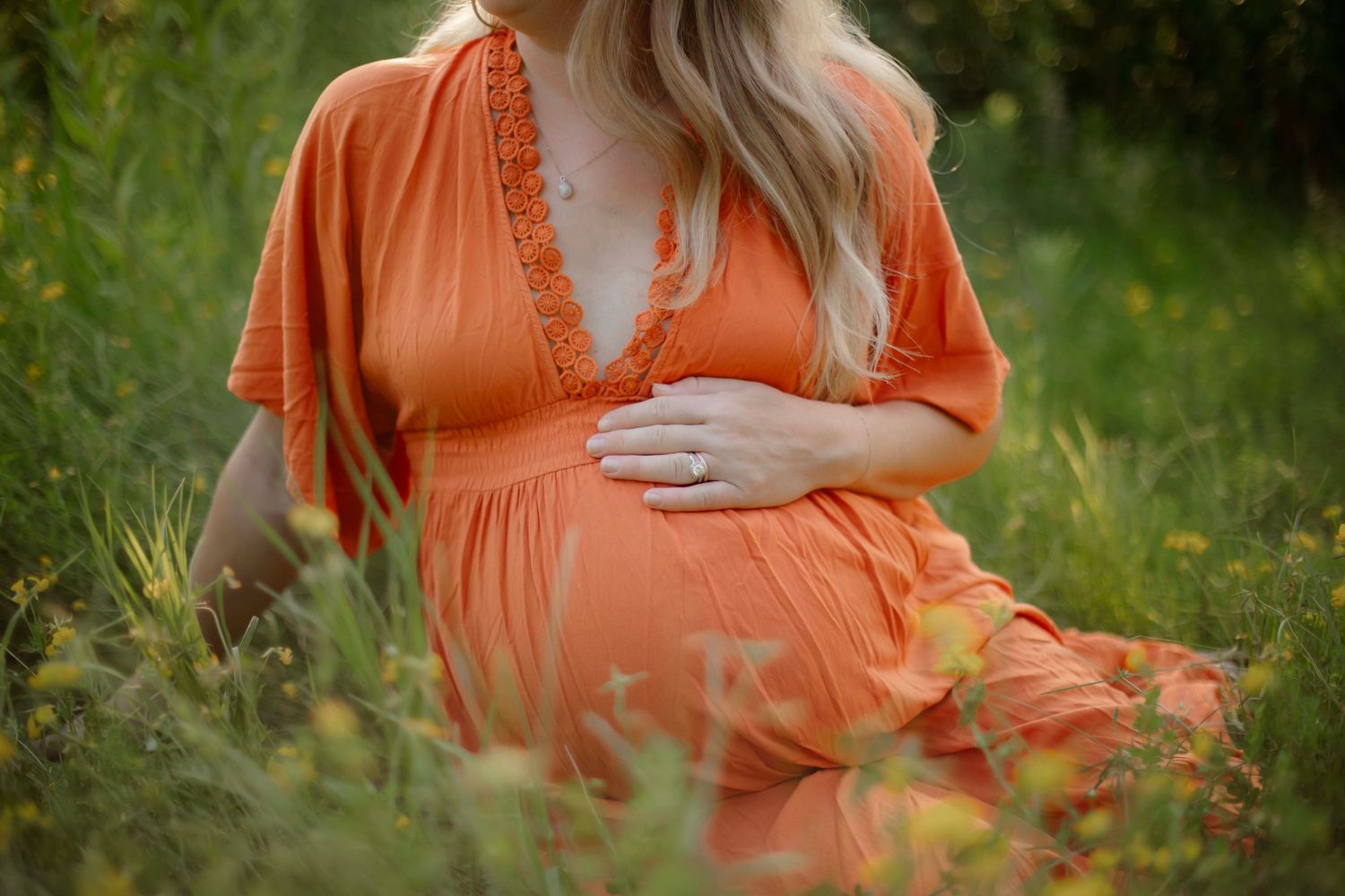 Pregnant woman in orange dress sitting in green field, cradling baby bump with one hand outdoors.