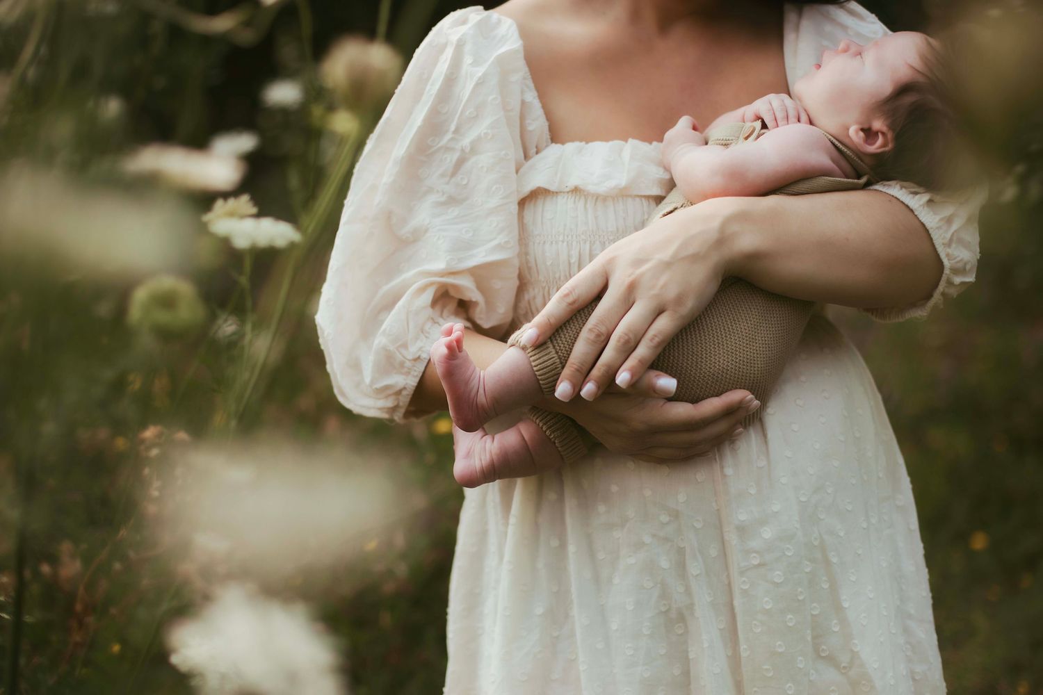 A mother in a white lace dress cradles her newborn baby in a sunlit wildflower meadow.