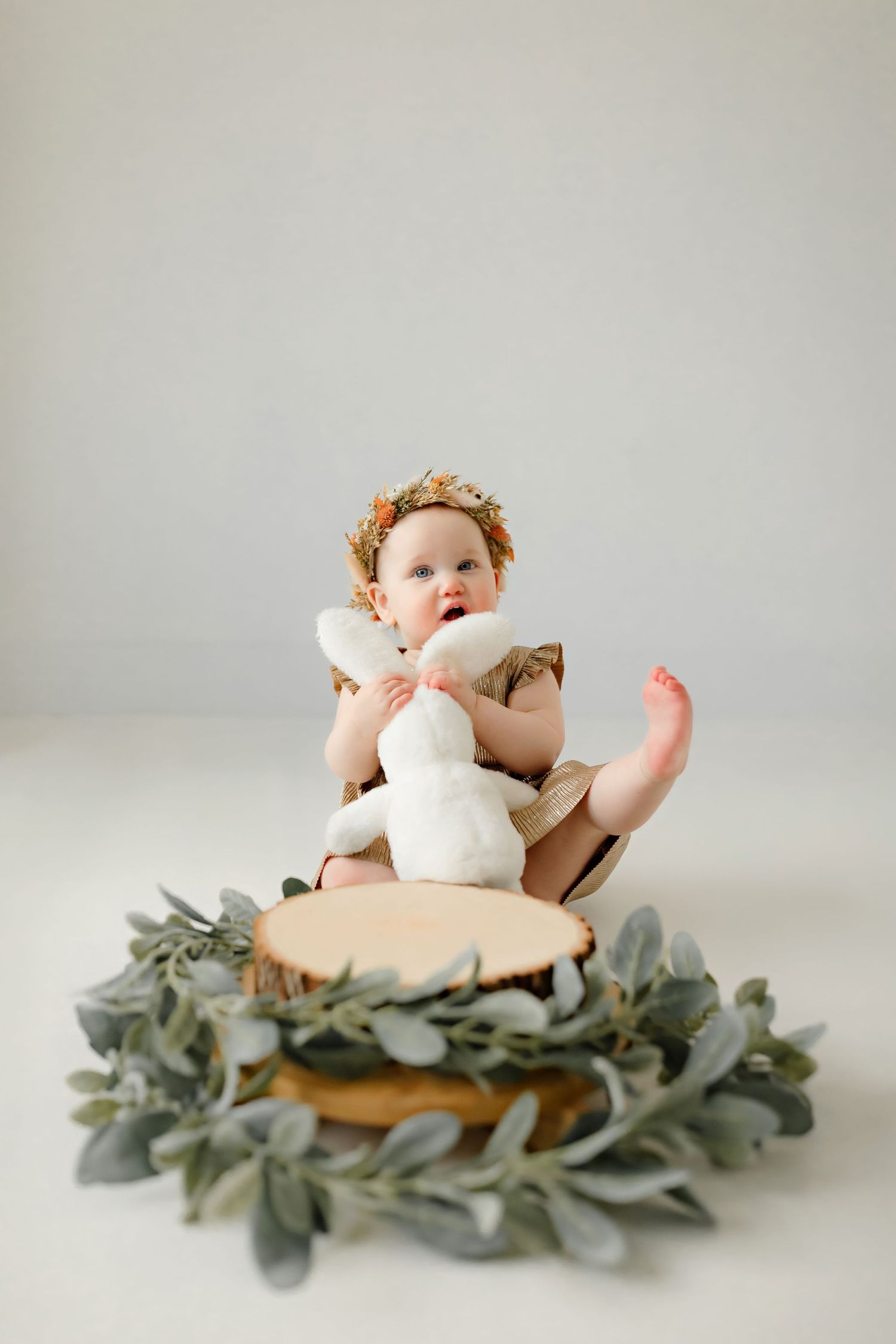 Baby wearing gold crown holding bunny toy on wooden disc surrounded by eucalyptus.