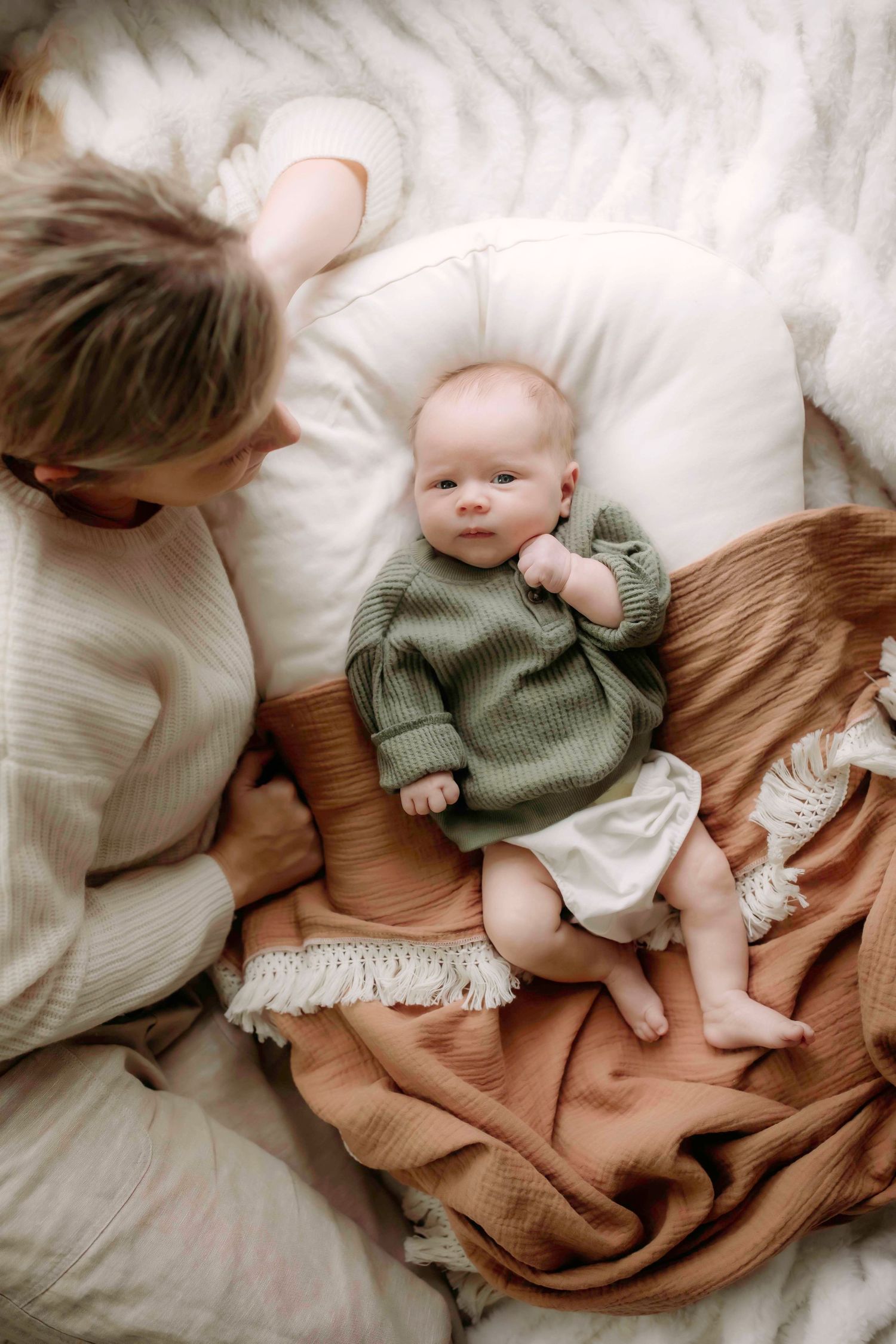 Baby in green sweater lying on brown blanket while caregiver looks on lovingly.