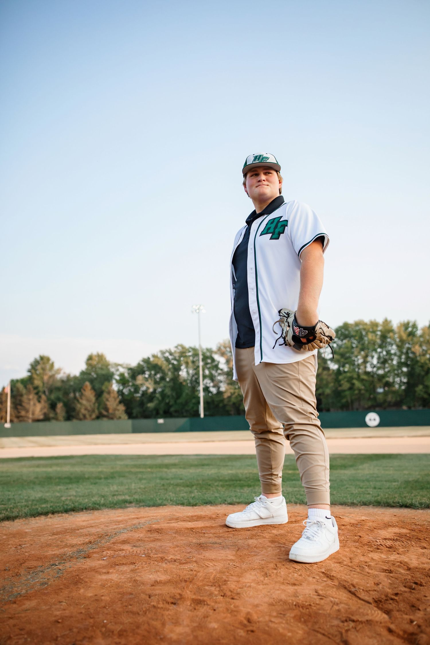 Baseball player poses on pitcher's mound in white jersey and khaki pants at baseball field.