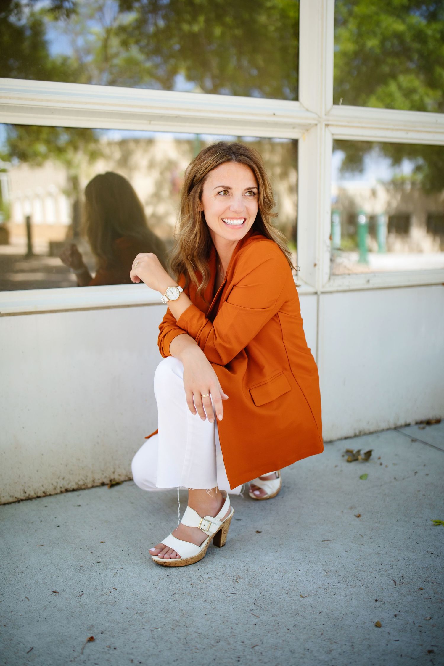 Woman in orange blazer and white pants crouches by window in casual pose.