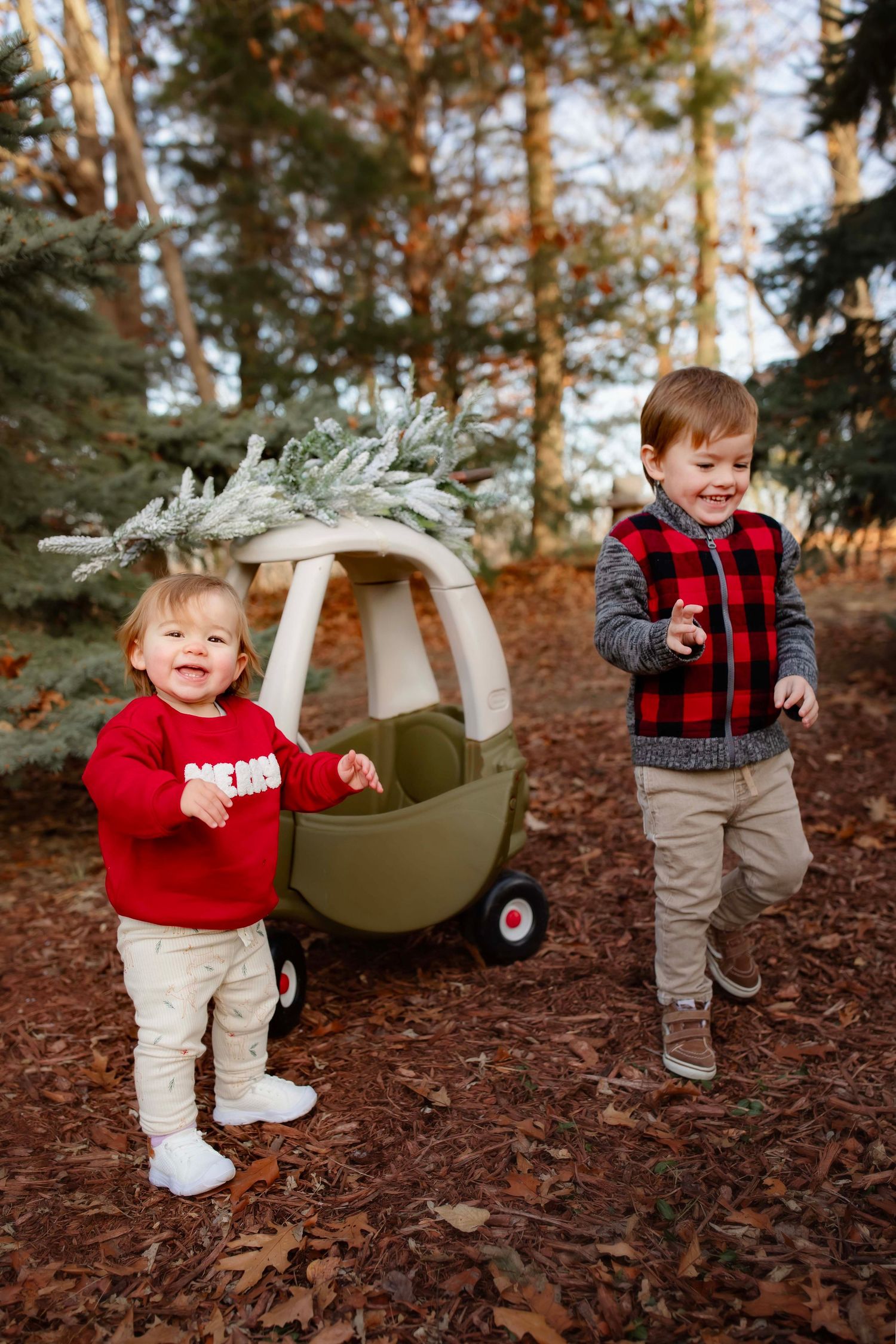 Two young children play outdoors with a decorated toy cozy coupe car in a wooded forest setting.