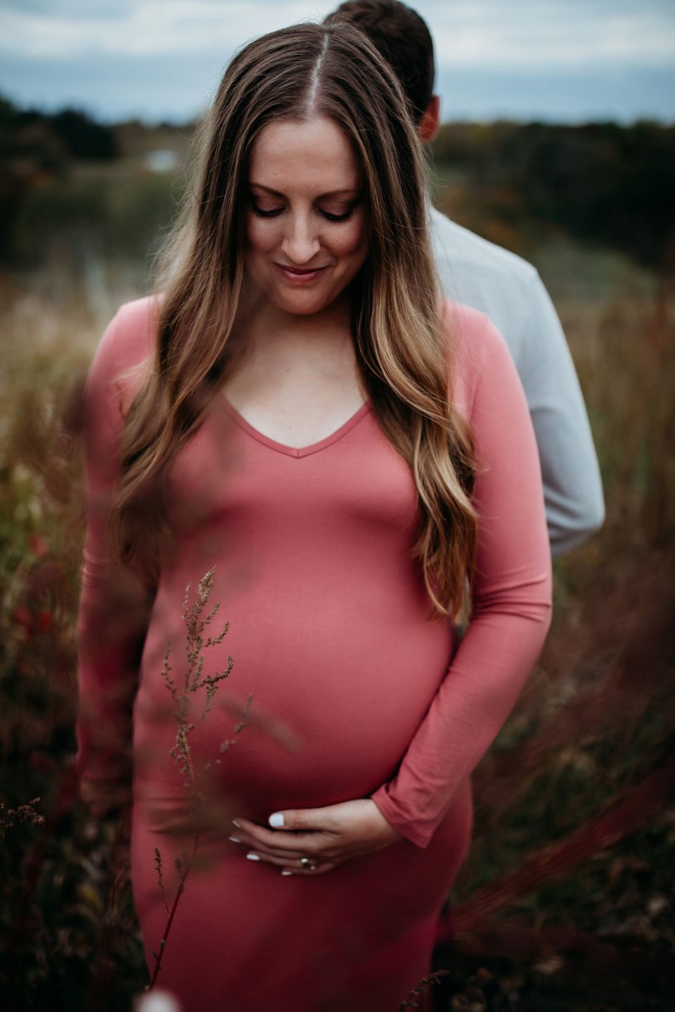 Maternity portrait of a woman in a pink dress touching her belly among wildflowers.