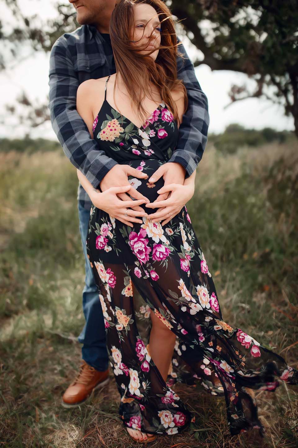 A couple embraces in a field while she wears a flowing black floral dress and he wears a plaid flannel shirt.