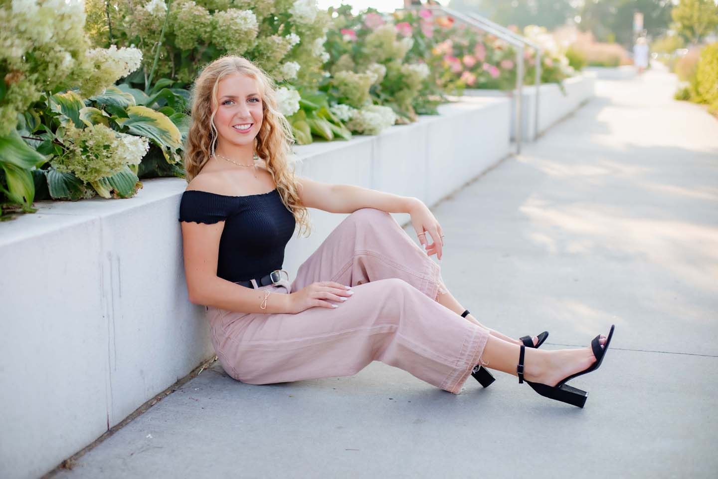 Casual outdoor pose on white wall wearing pink pants and black top with heels.