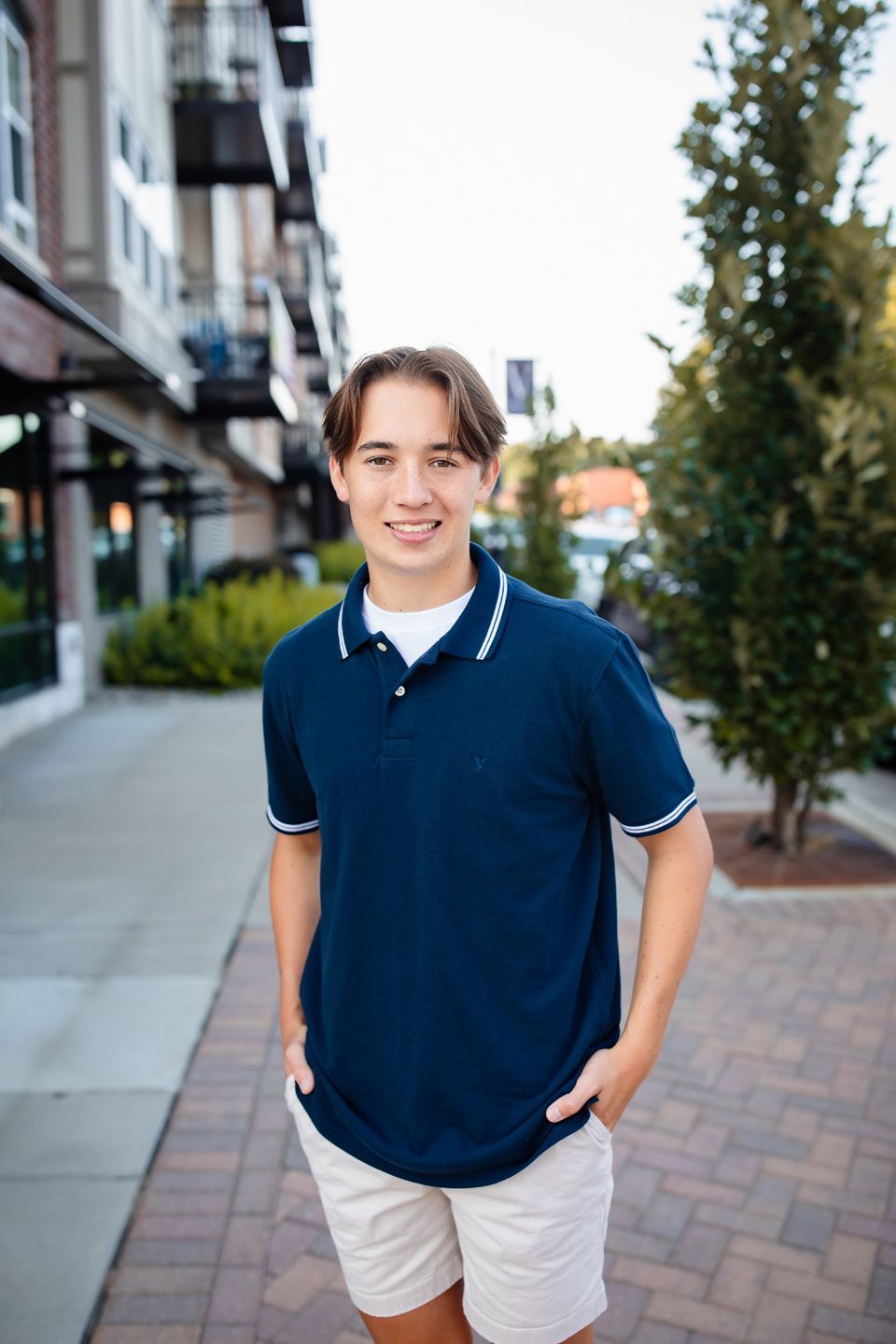Someone in a navy blue polo shirt and white shorts stands smiling on a brick sidewalk near apartment buildings.