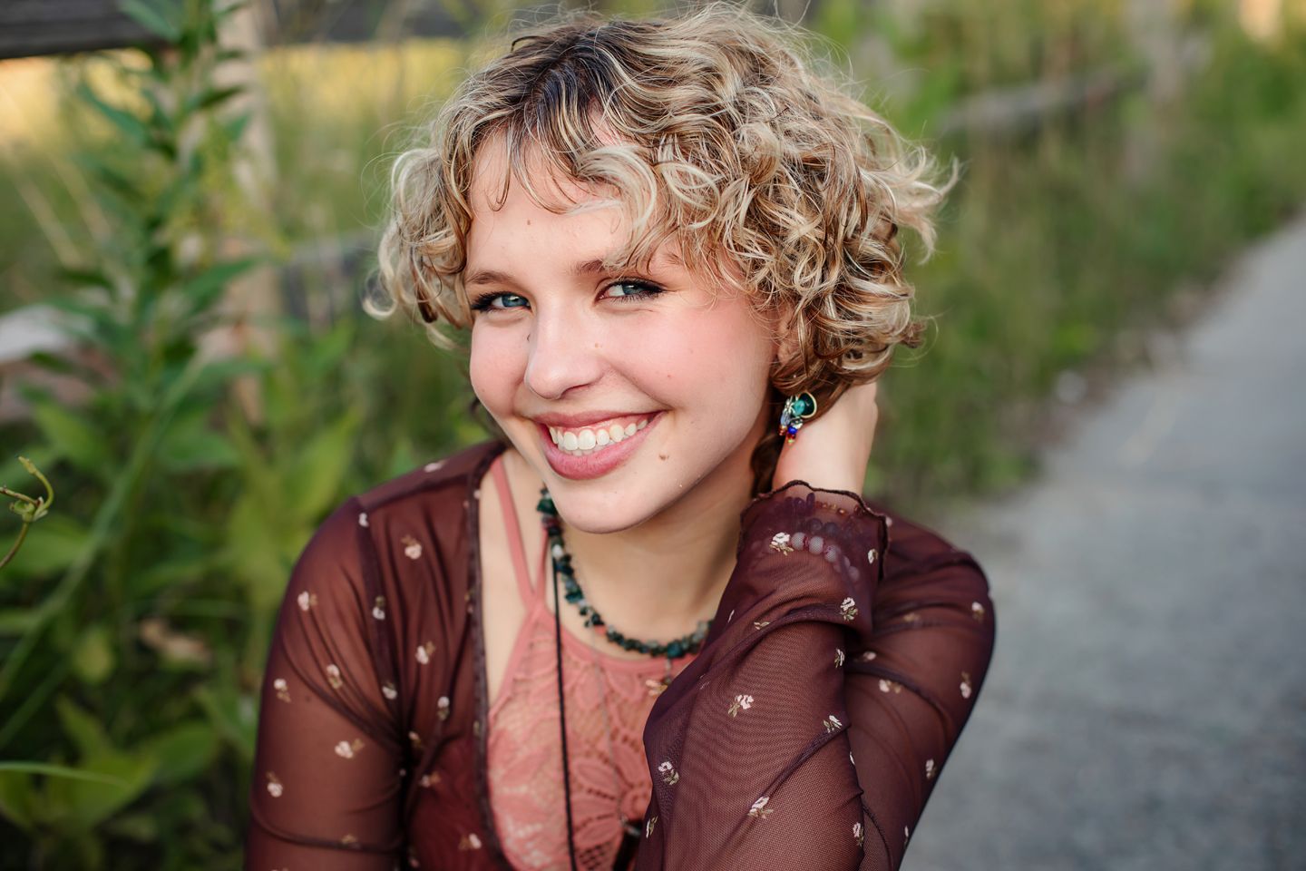A warm outdoor portrait with a sheer brown polka dot top and curly blonde hair against a soft natural background.
