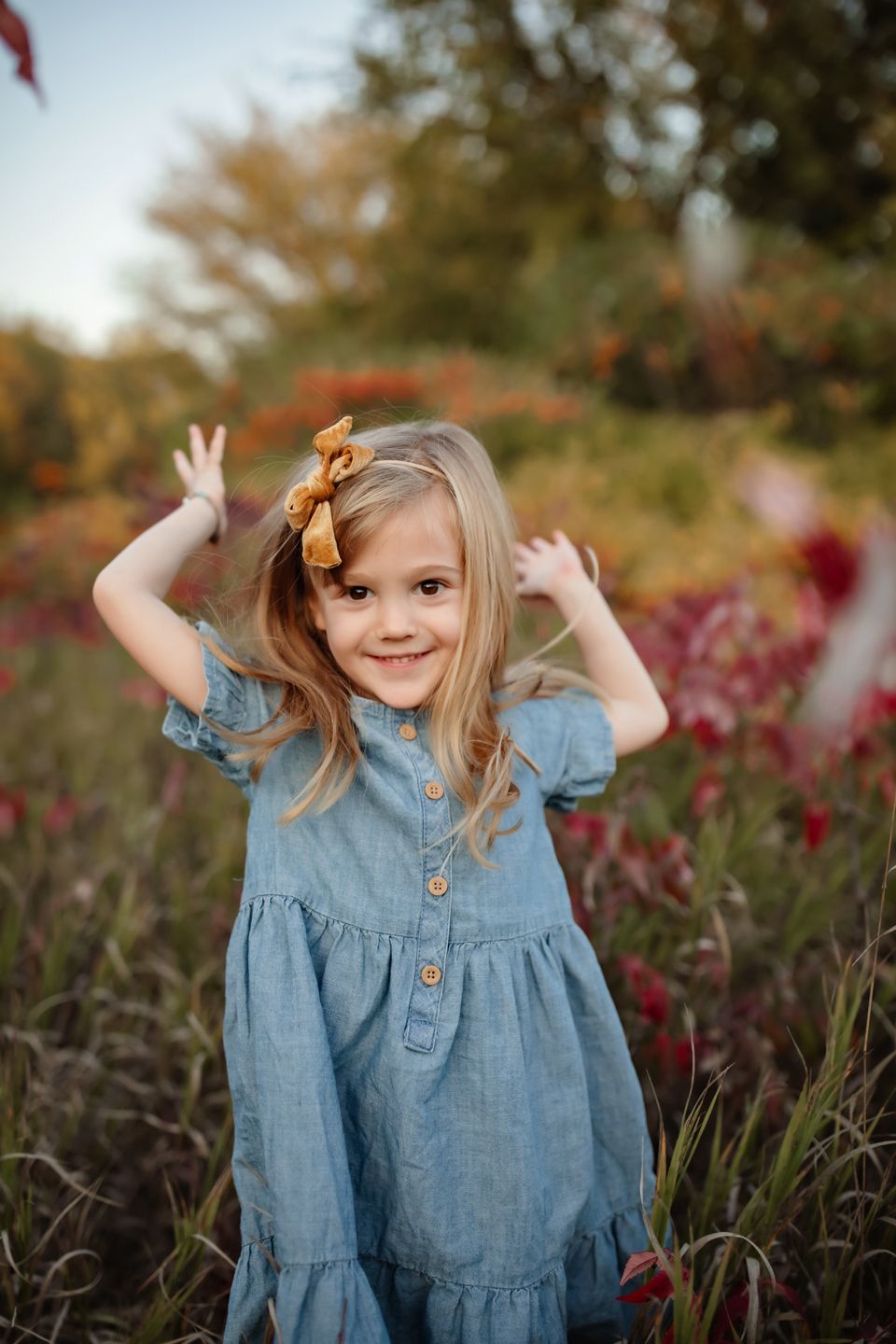 Young child in blue denim dress with yellow bow raising hands playfully in autumn field.