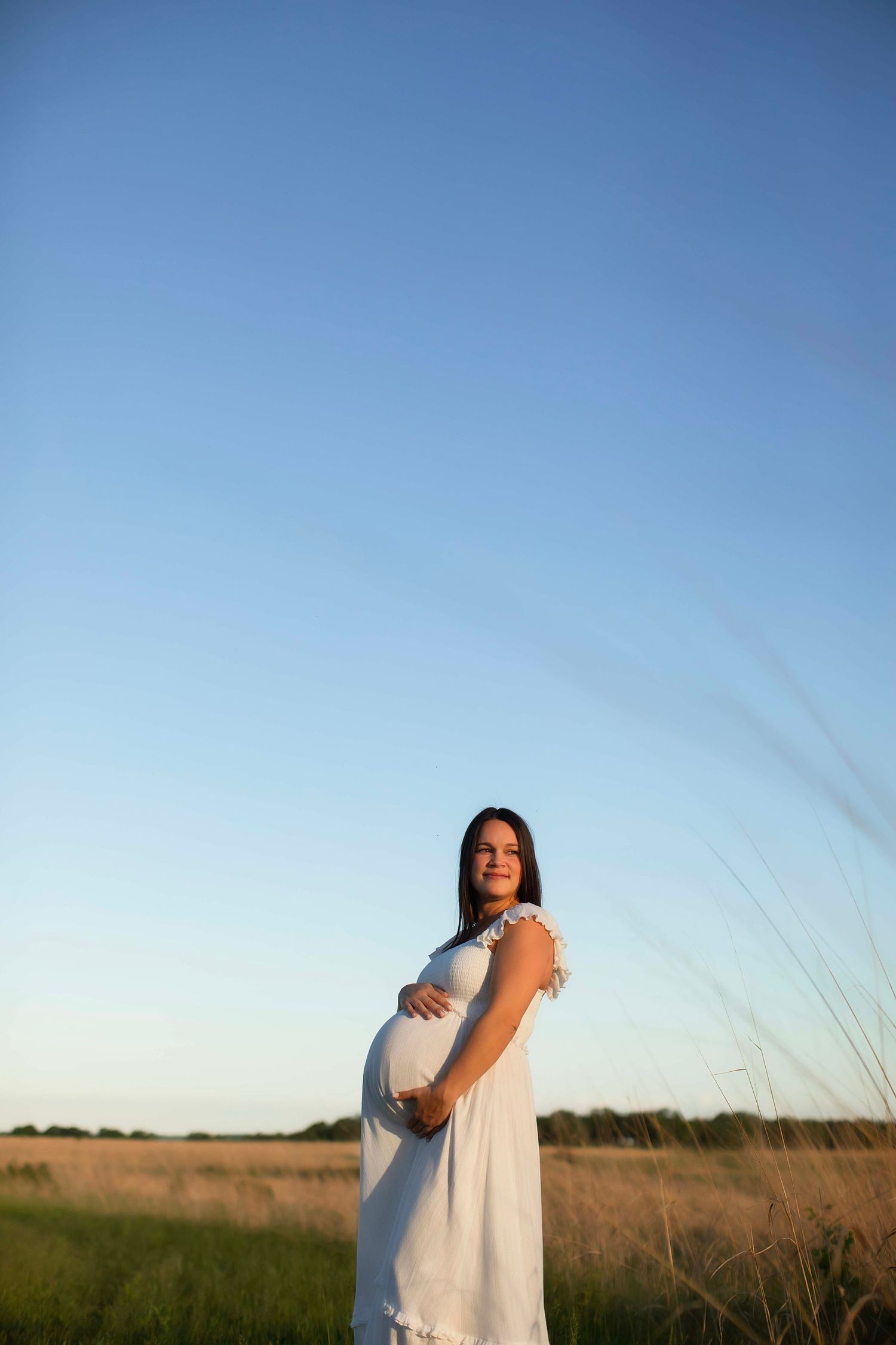 Pregnant woman in white dress standing in golden field under clear blue sky, cradling baby bump at sunset.