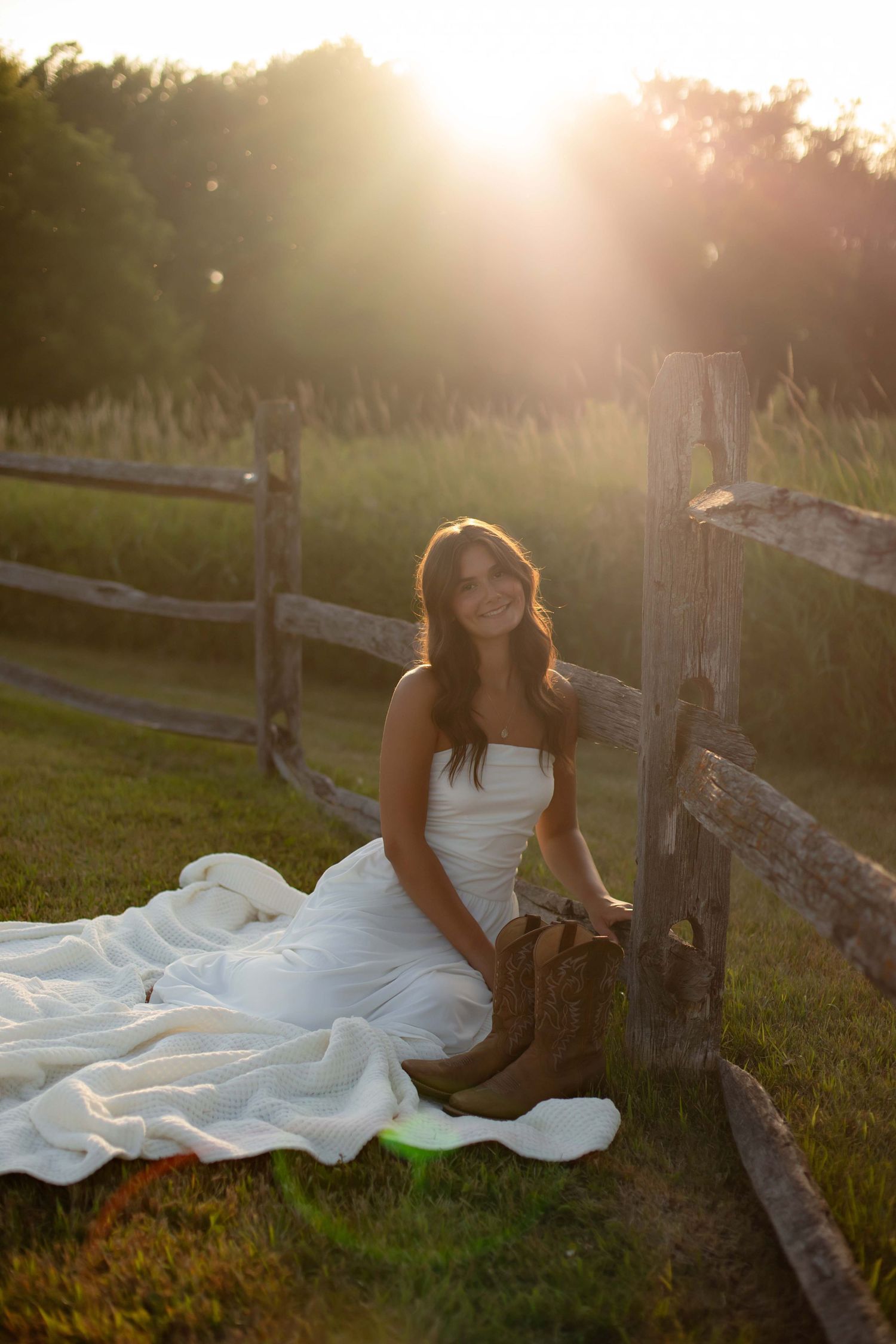 Bride in white strapless gown and cowboy boots sitting by a rustic fence at golden hour sunset.