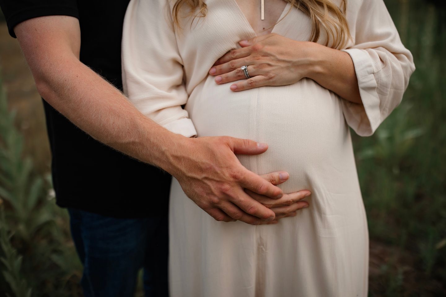 Close-up shot of hands tenderly cradling a pregnant belly in a white maternity dress against a natural backdrop.