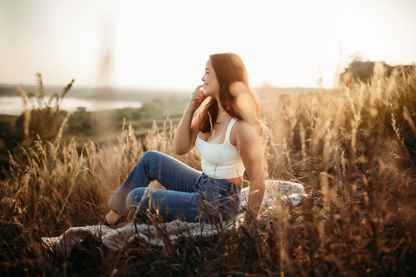 Woman sitting peacefully in tall grass during golden hour with scenic landscape behind.