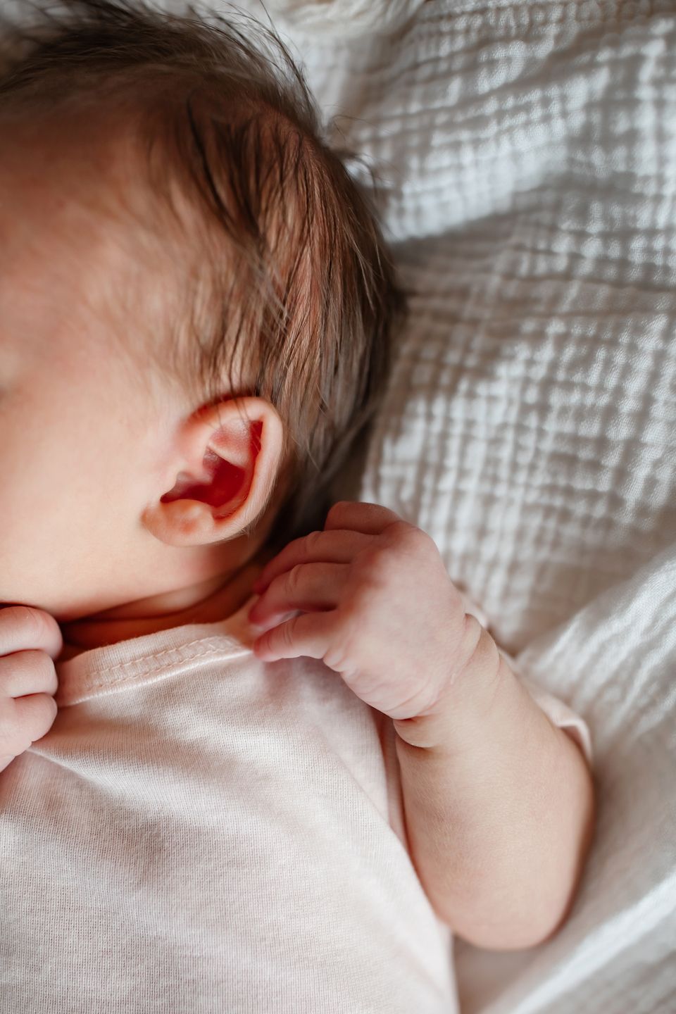 Close-up of baby's ear and tiny hand against soft white bedding.