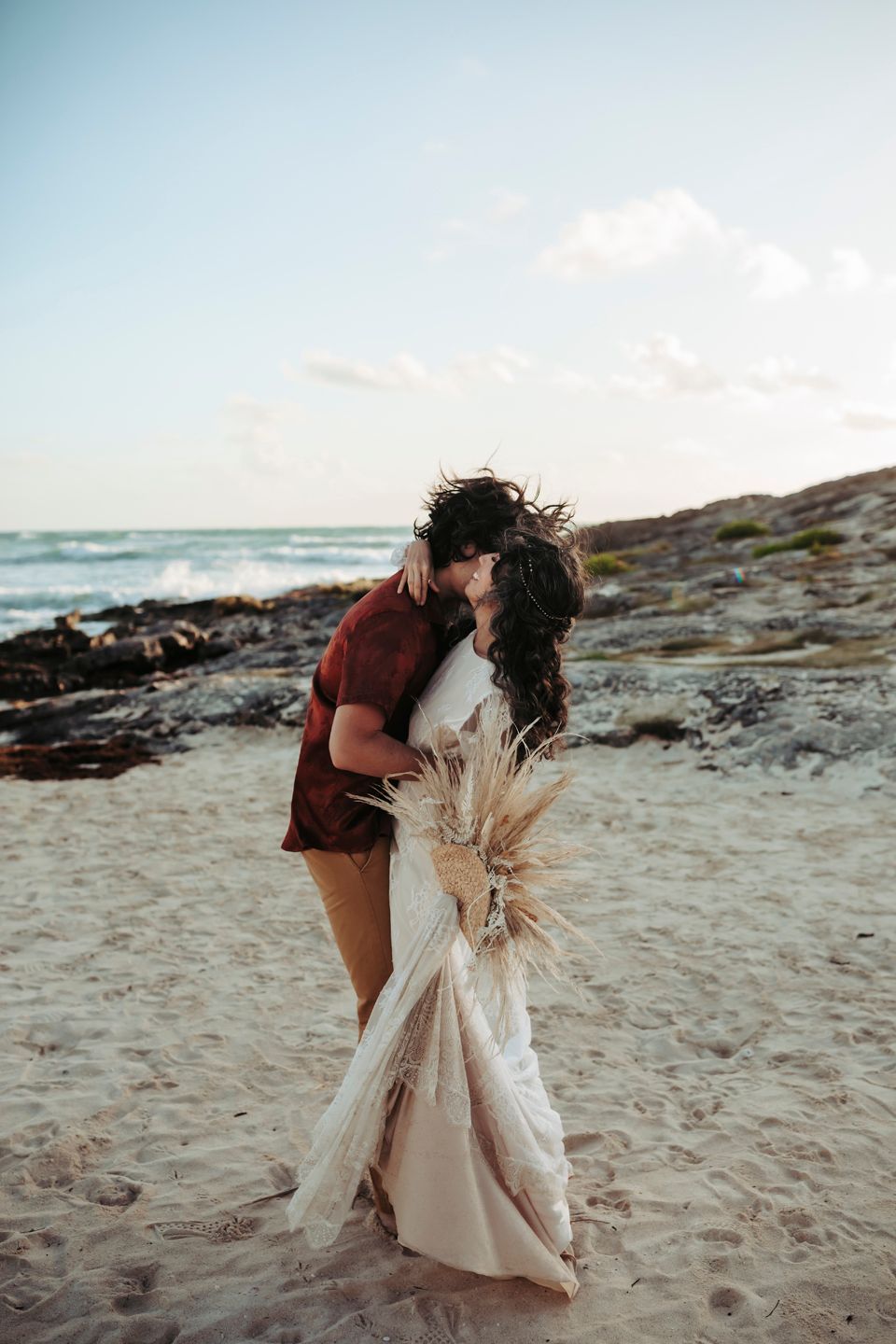 Romantic beach moment with white dress and dried grass bouquet against coastal rocks.