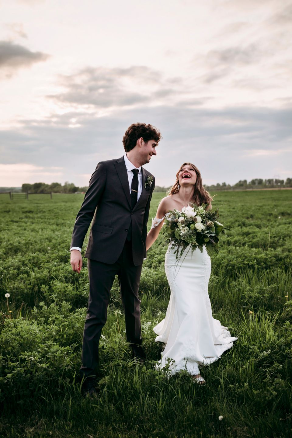 Joyful newlyweds share laughter while standing in midwest verdant field at sunset.