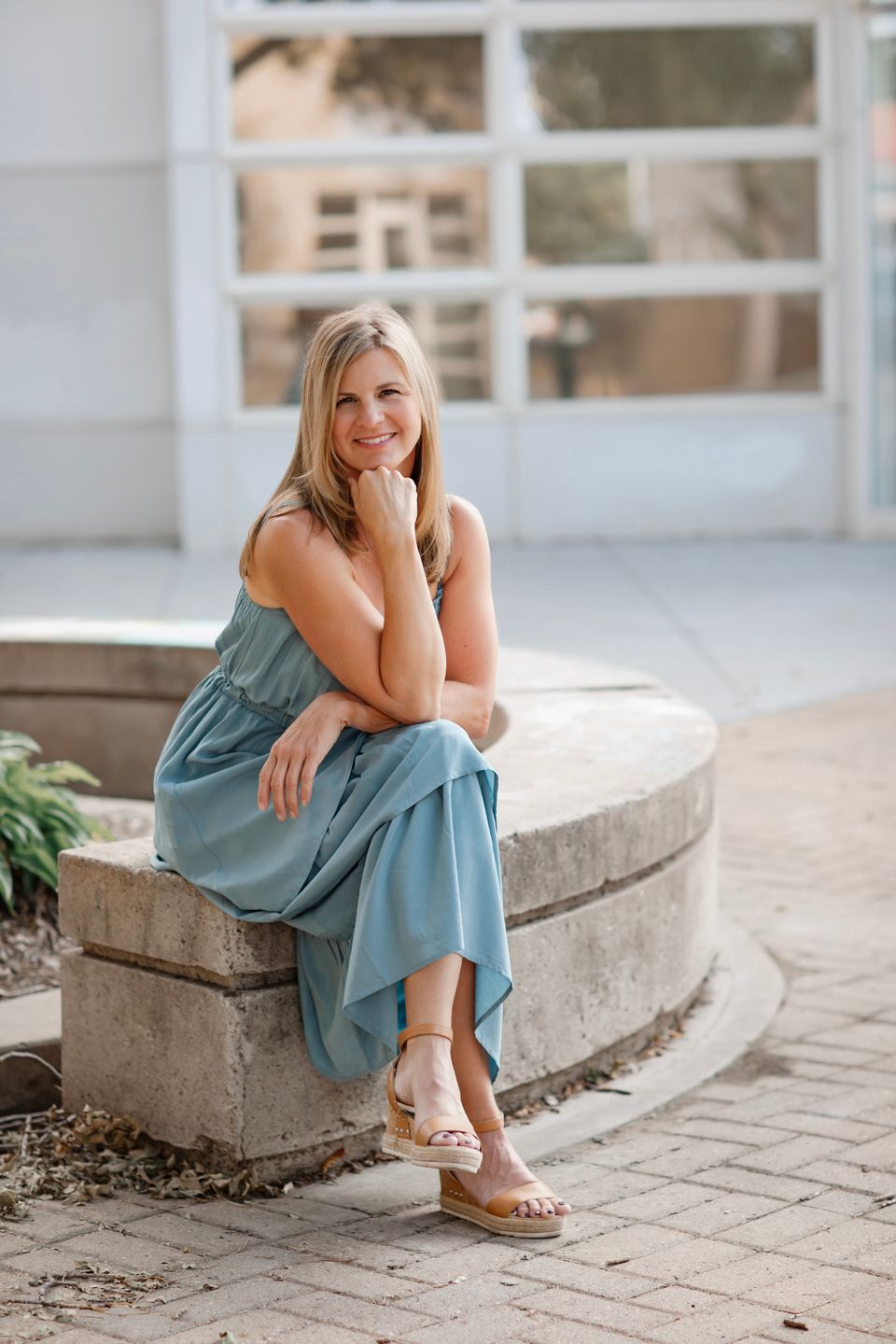 Lifestyle portrait sitting on concrete ledge wearing casual blue dress and sandals.