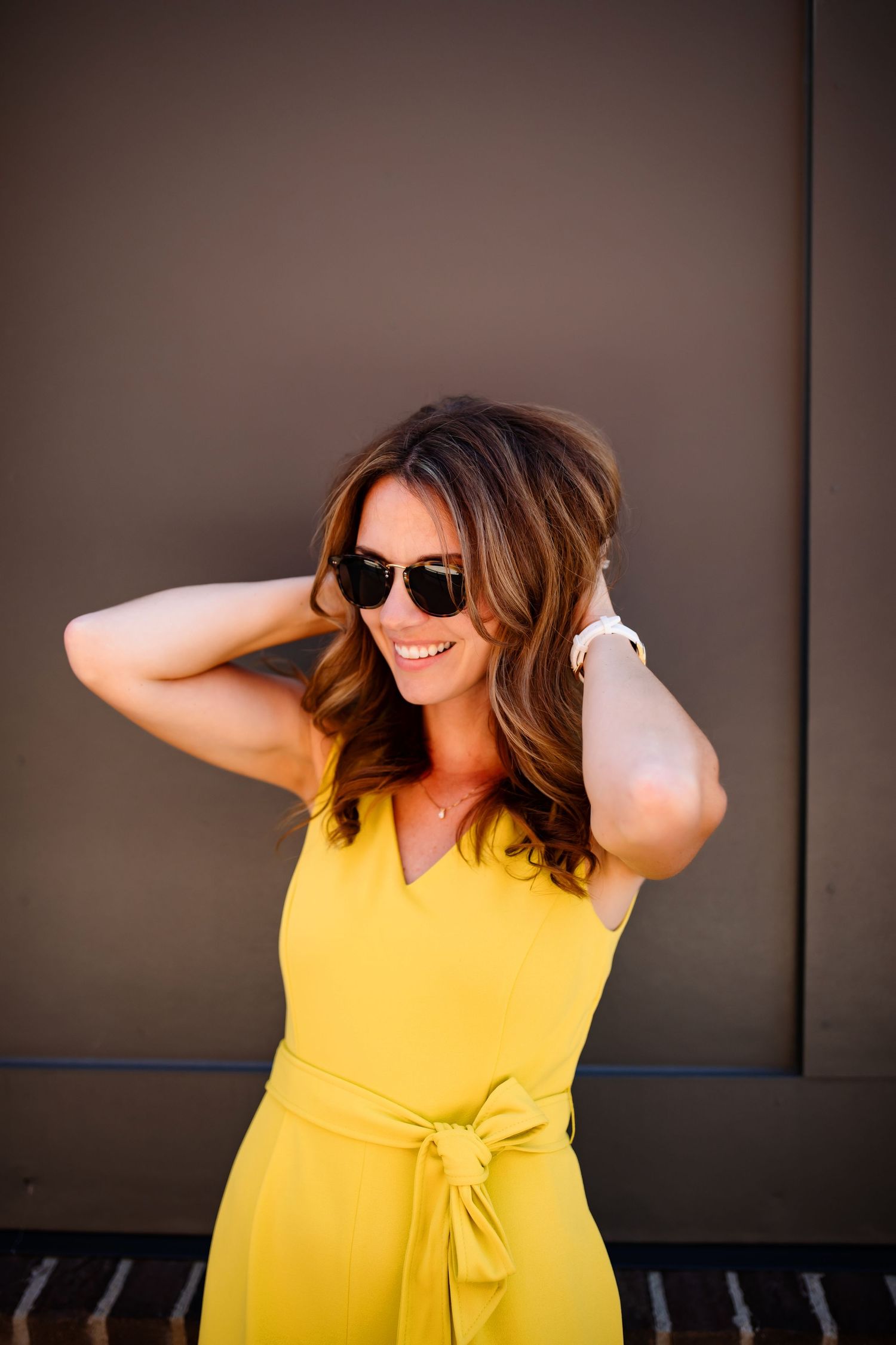 Fashionable woman in yellow dress and sunglasses poses against dark wall with joyful expression.