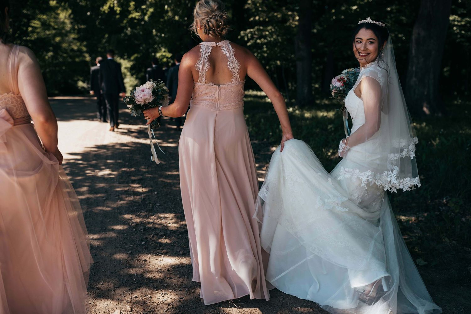 Bridesmaids in blush gowns assist the bride with her dress in a sunlit wooded setting.