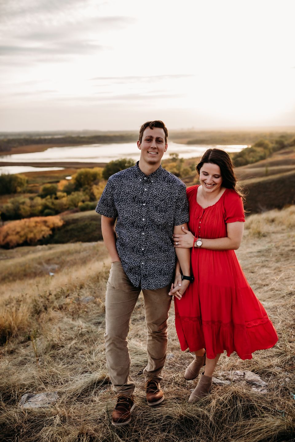 A couple poses together on a hillside wearing coordinating red dress and navy patterned shirt.