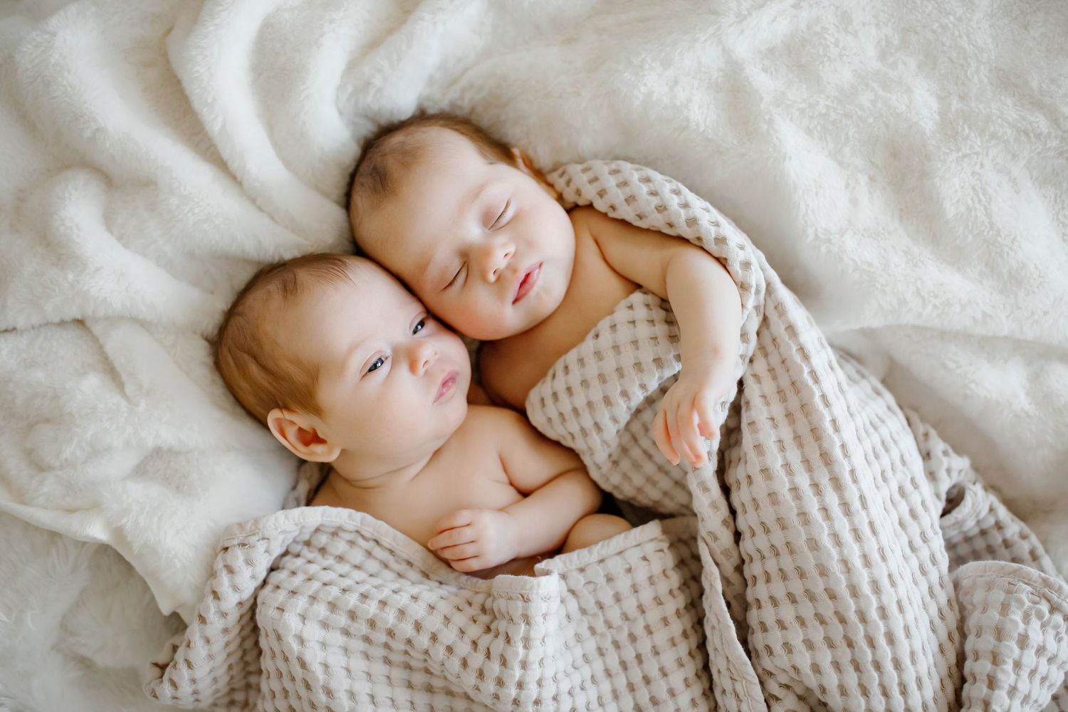 Twin babies sleeping peacefully together wrapped in textured grey blanket.