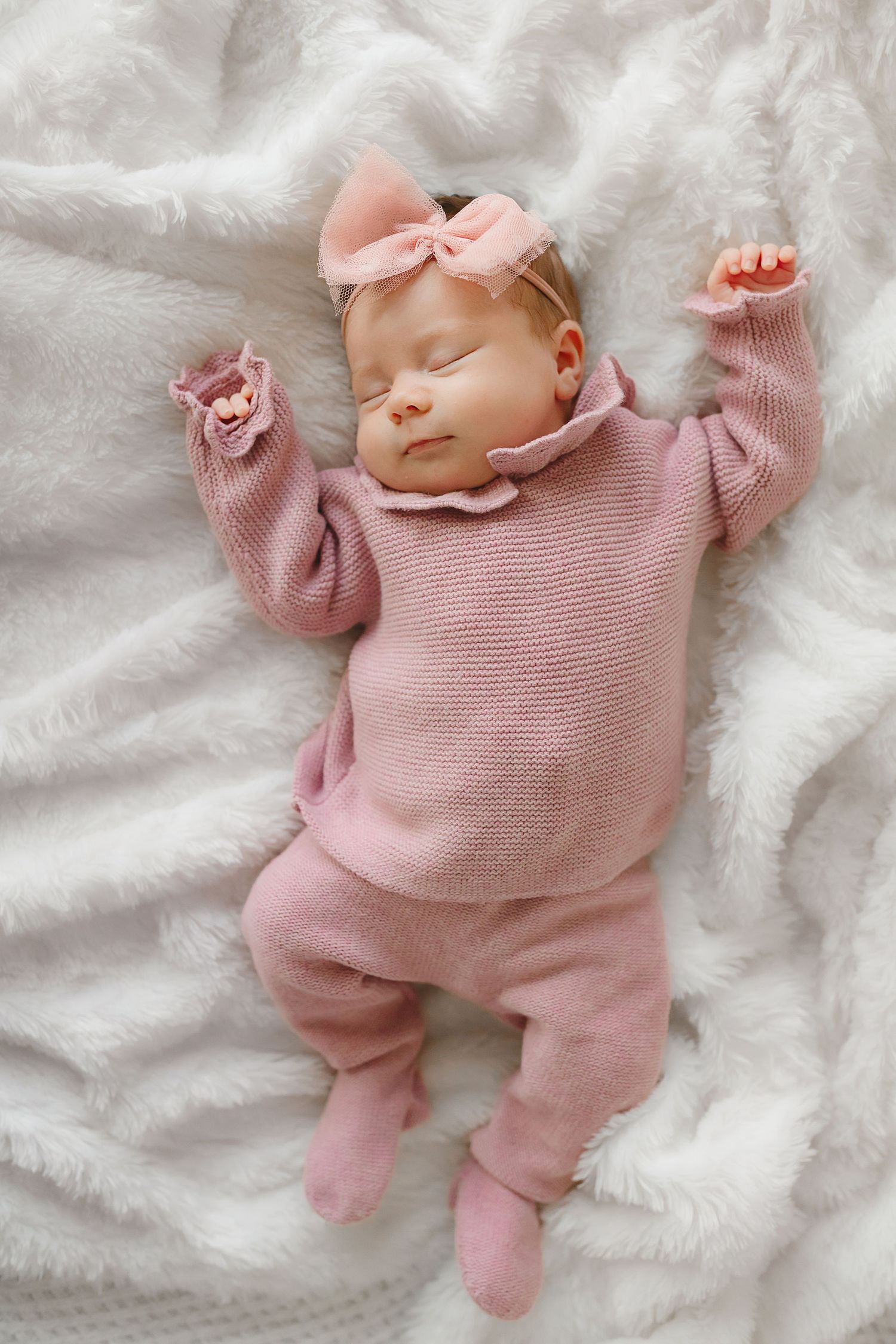 Sleeping baby in pink knit outfit with matching bow on white bedding.