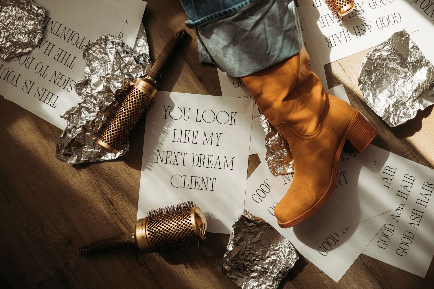 Flat lay of brown heeled boots, crumpled foil, and papers reading 'You Look Like My Next Dream Client'. Hair Stylist branding shoot.
