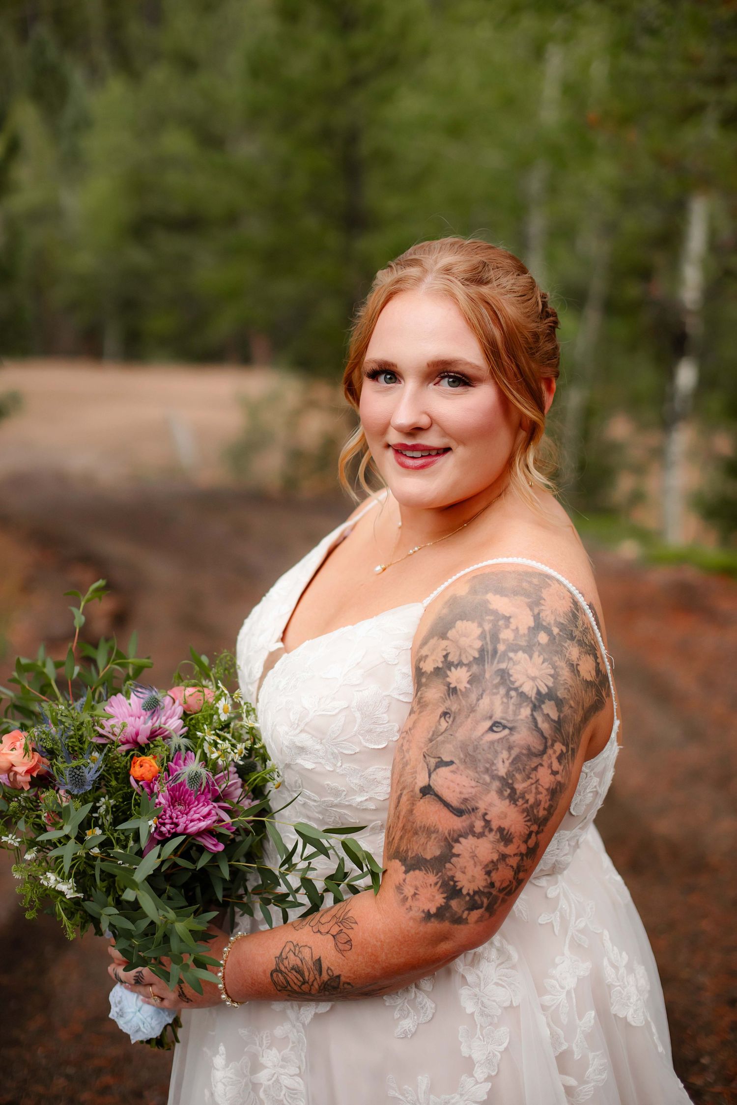 A person in a white wedding dress with floral lace details holds a colorful bouquet outdoors among trees.