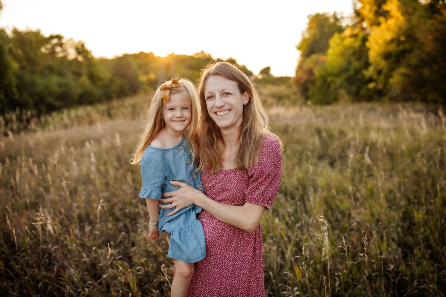 Mother and daughter embrace in a golden sunlit meadow at sunset wearing blue and burgundy dresses.