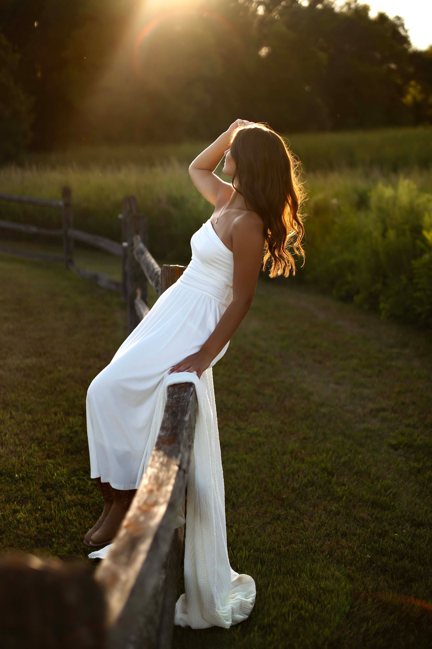 Woman in white dress and cowboy boots sits on rustic fence at golden sunset in lush green field.