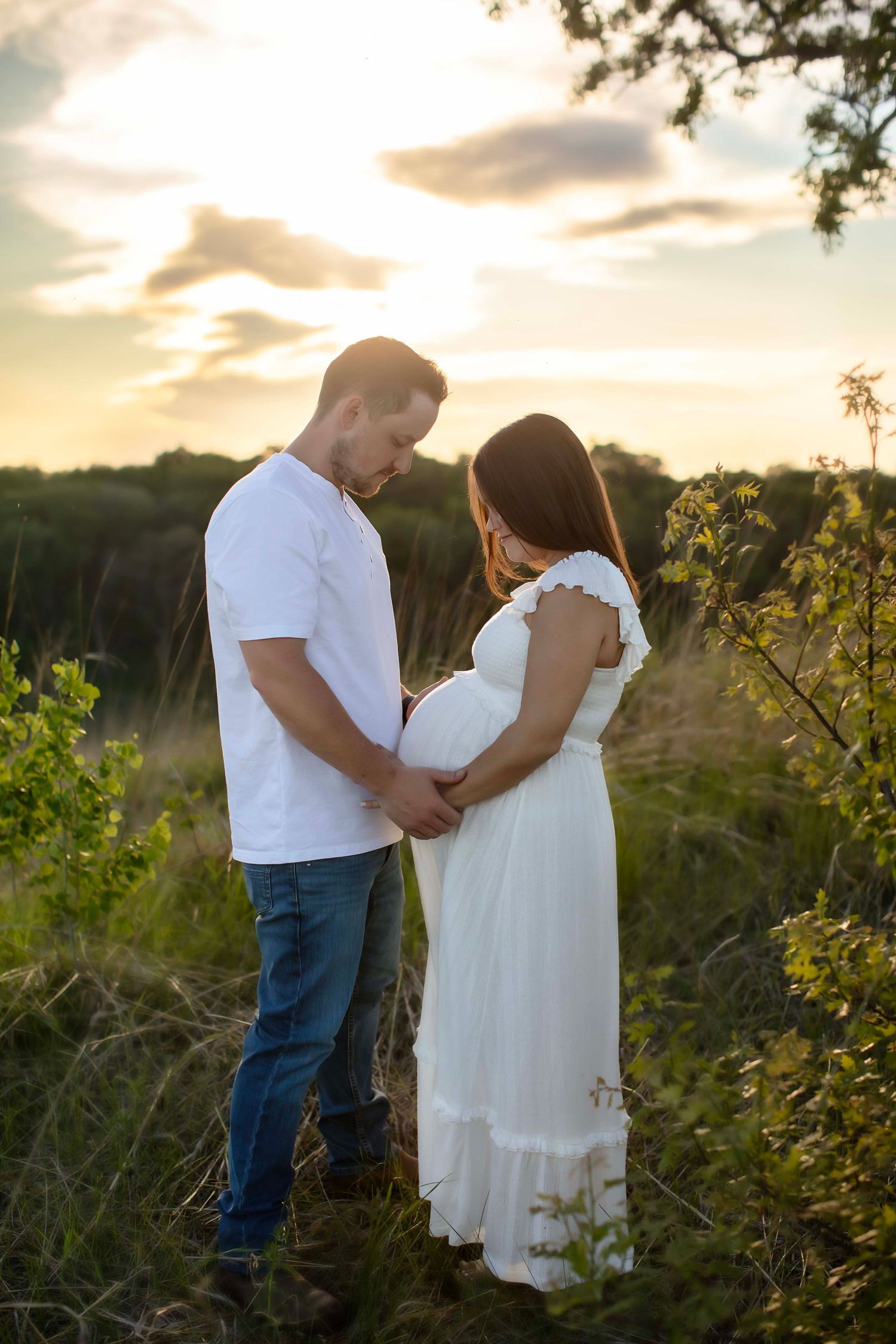 Couple holding pregnant belly together in golden sunset field outdoors.