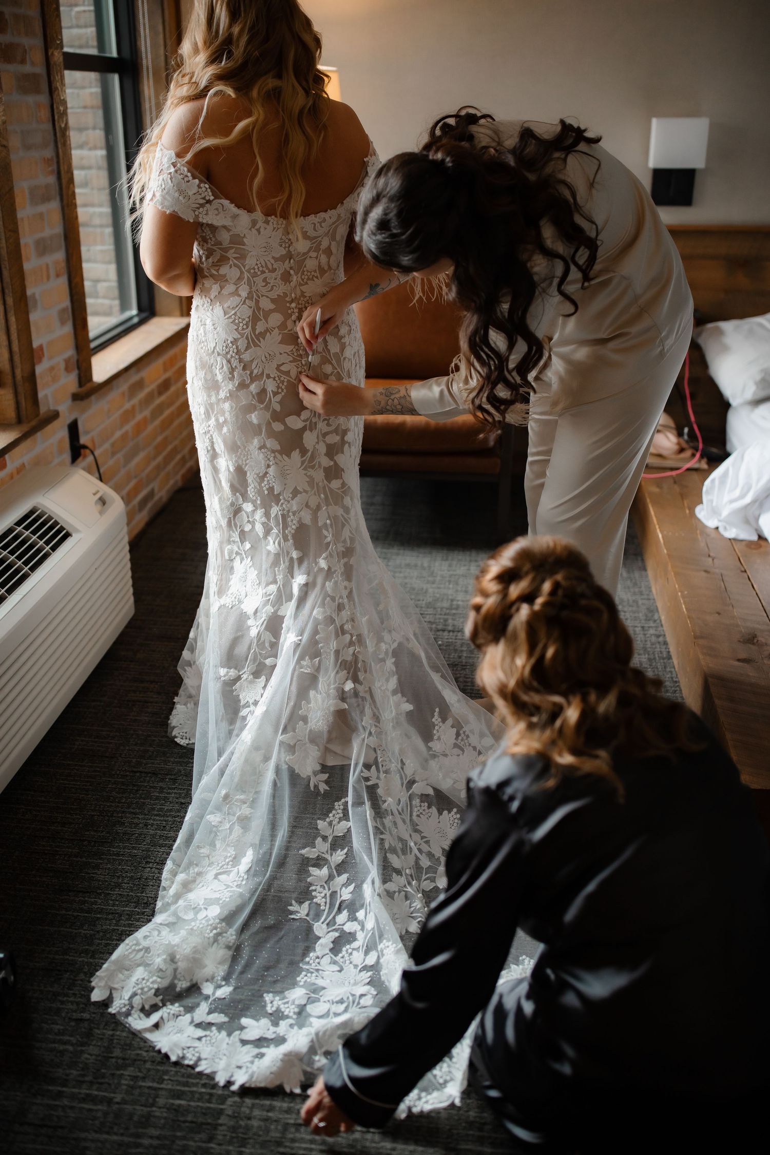 A bride in a detailed lace wedding gown being helped with the train and final dress adjustments in a rustic room.