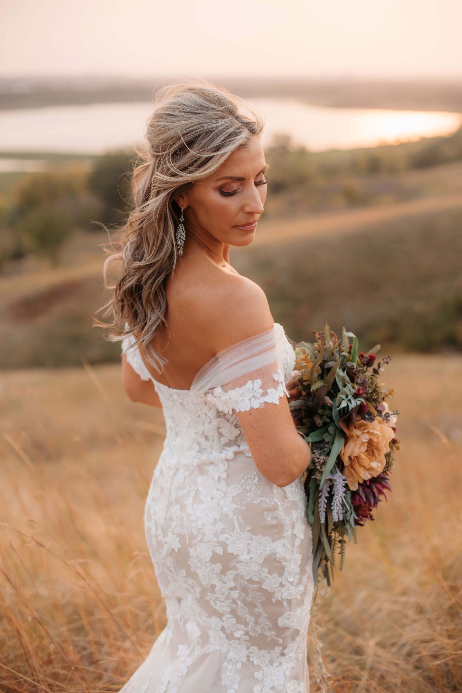 A bride in a lace off-shoulder gown poses with her bouquet in golden evening light.