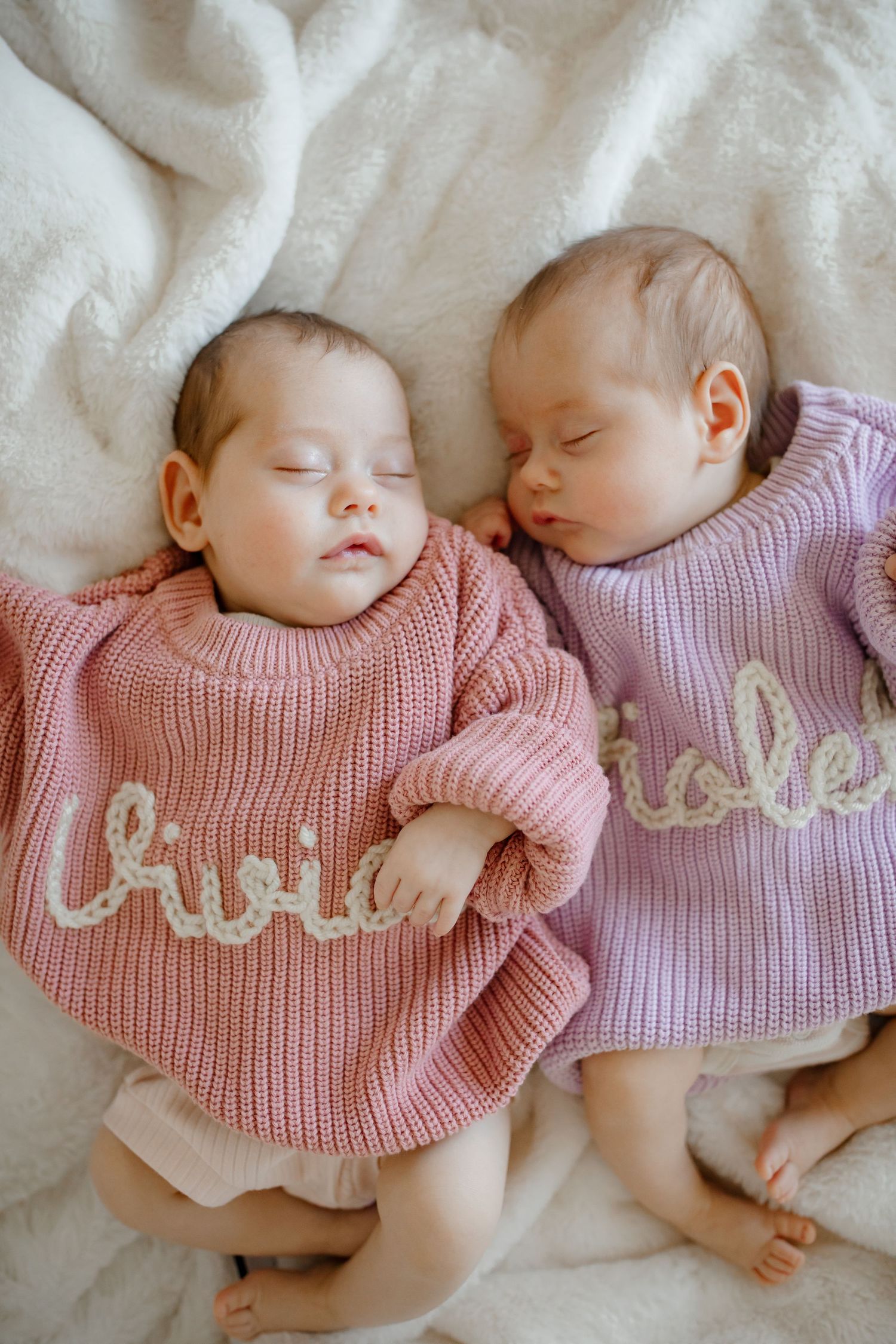 Twin babies in coordinating pink and purple knit sweaters sleeping on white blanket.