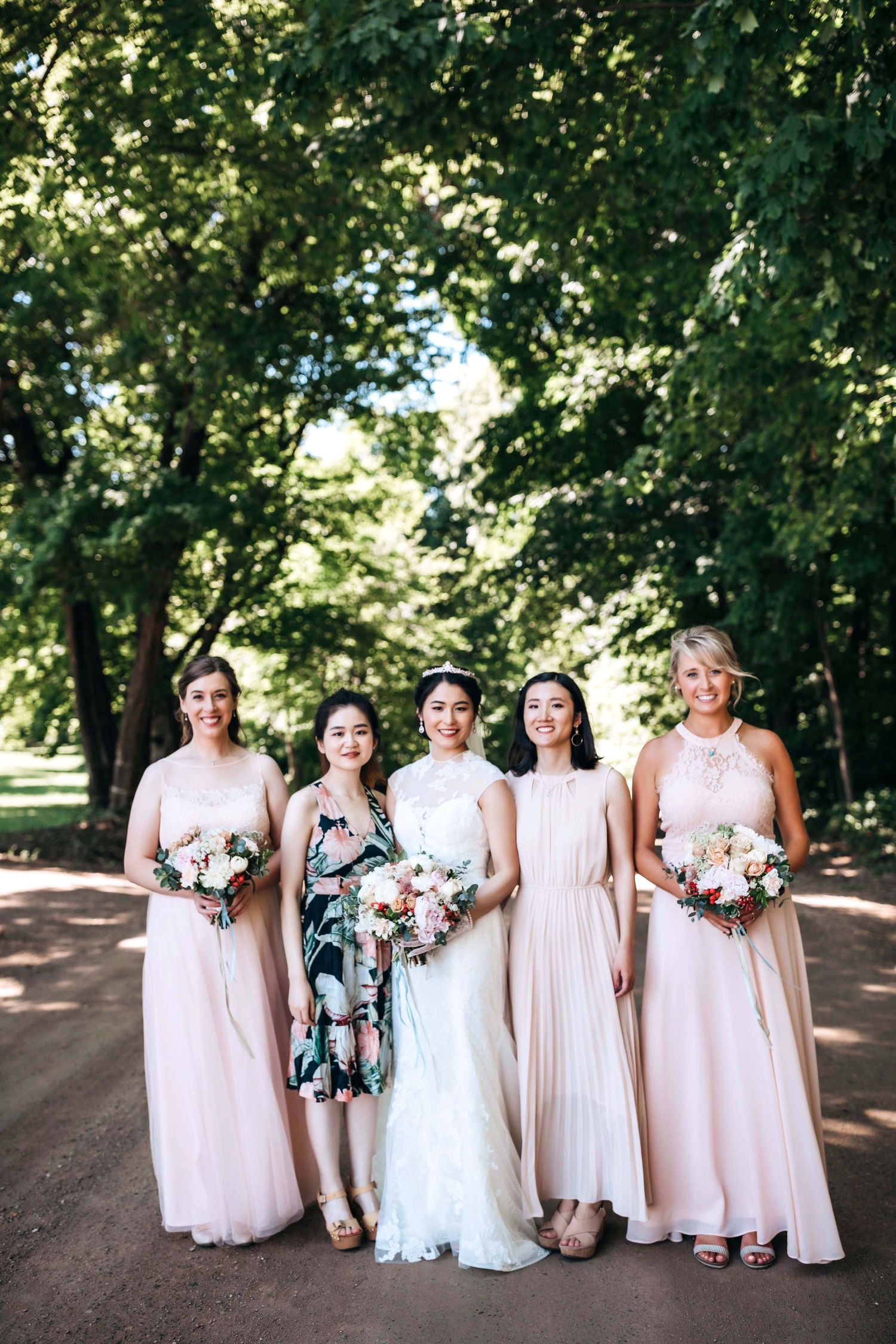 Bridal party of five women pose on a tree-lined path in coordinating pink and floral dresses.