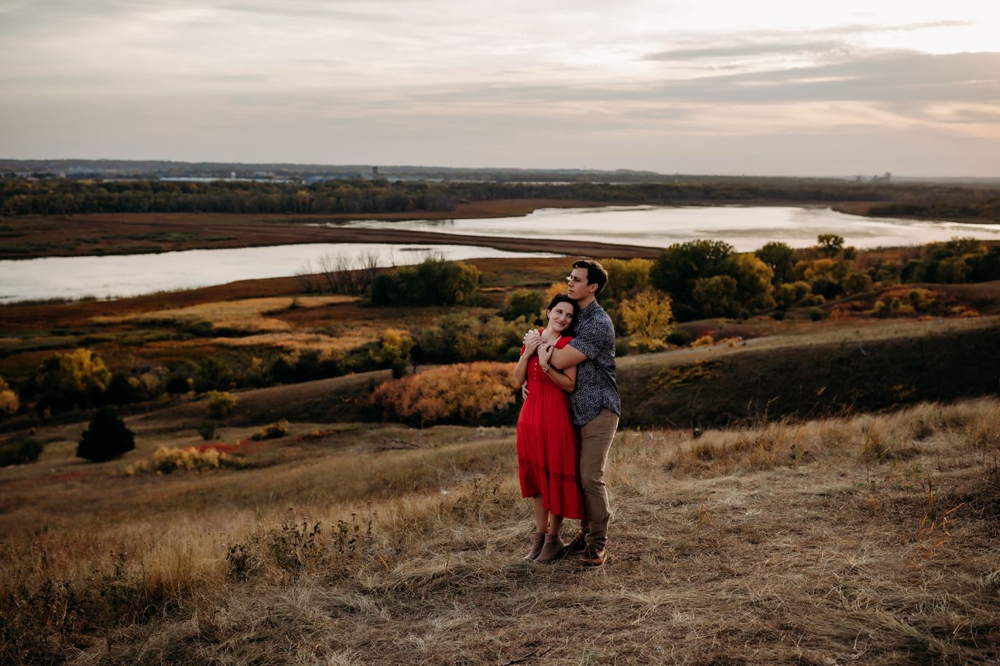 A couple embraces on a hilltop overlooking a lake during sunset.