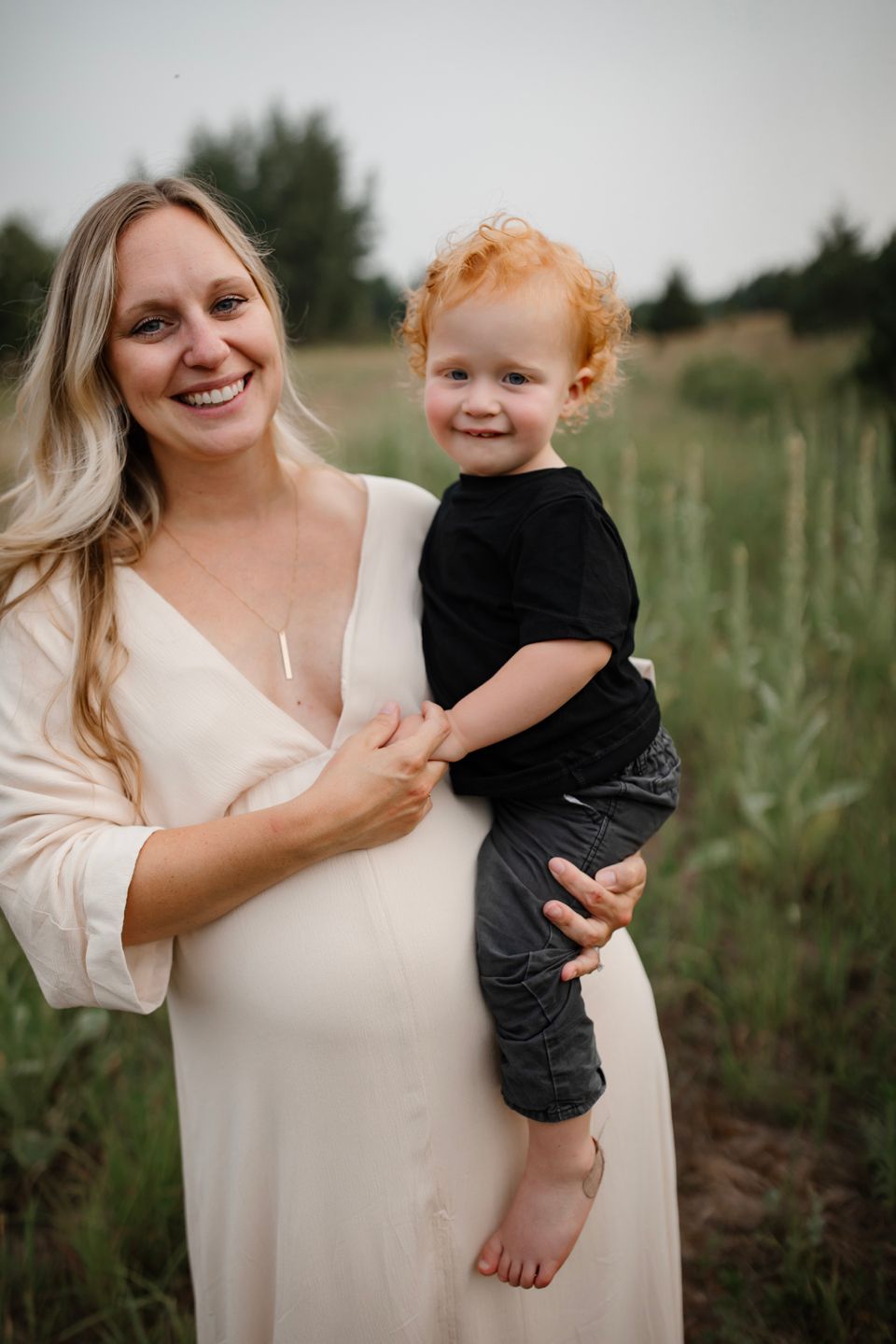 A maternity portrait shows an expectant mother in white holding a smiling toddler with red curly hair outdoors.