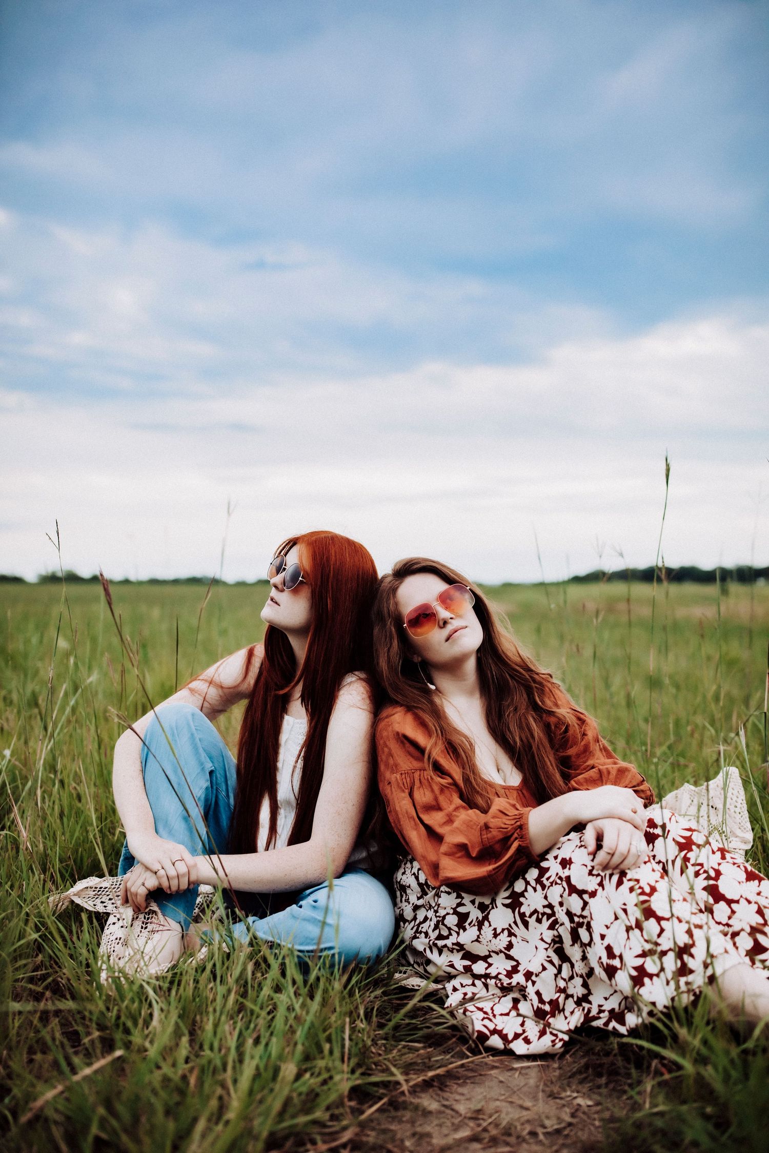 Friends in boho clothing sitting back-to-back in a grassy field under blue sky.