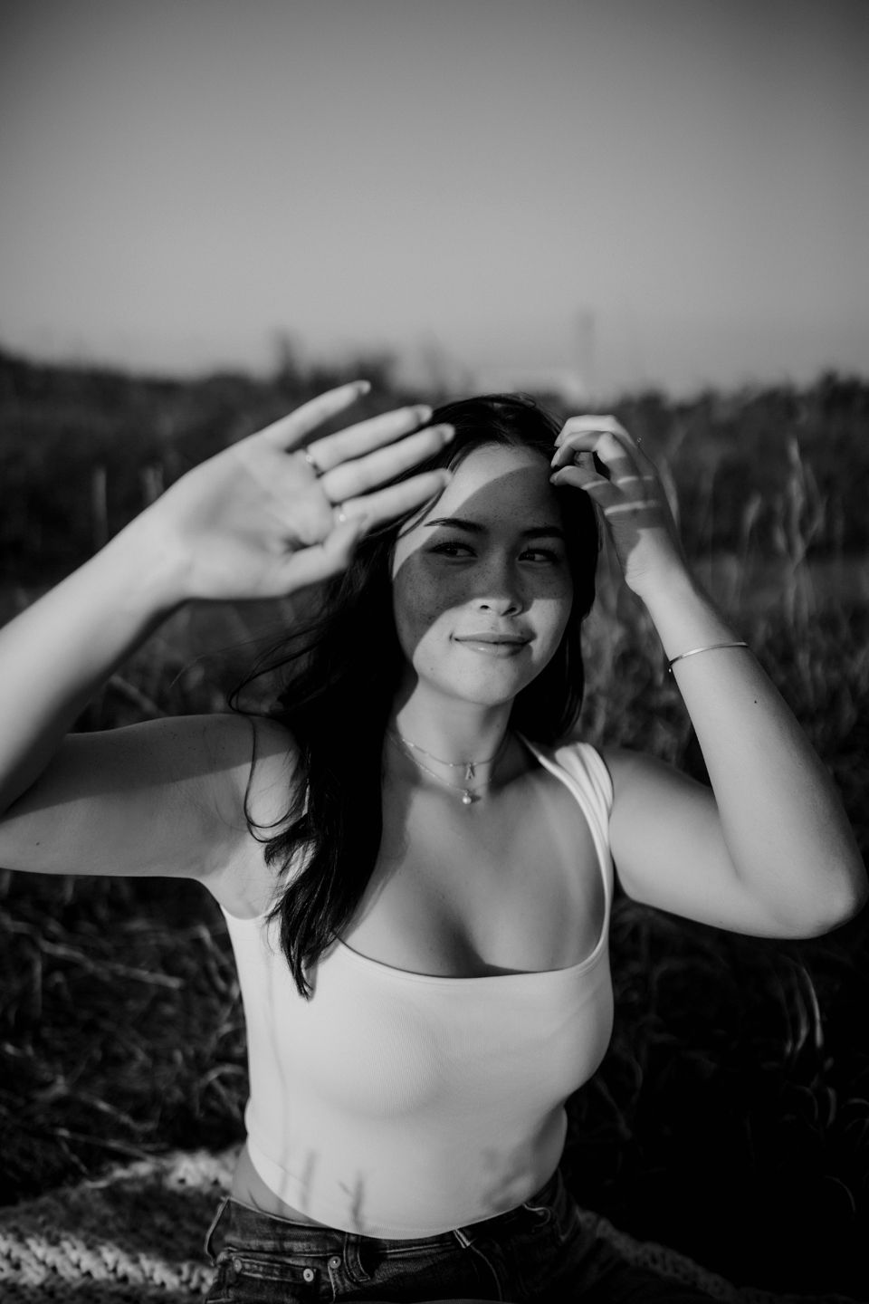 Black and white portrait of someone in a white tank top shielding their eyes from sunlight in a natural outdoor setting.