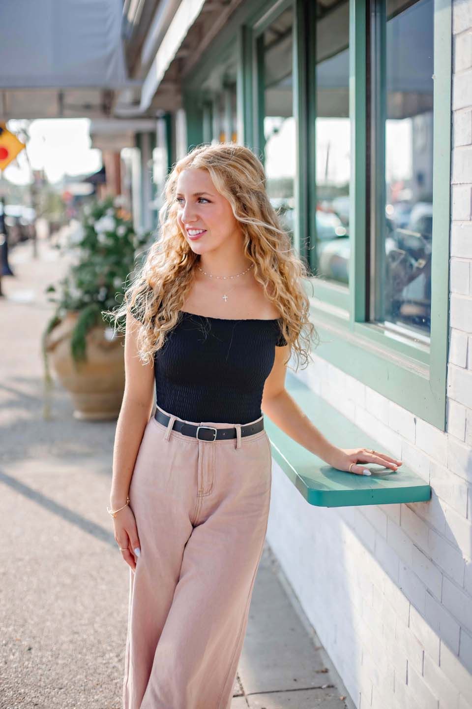 Person in black top and pink pants poses against storefront window.