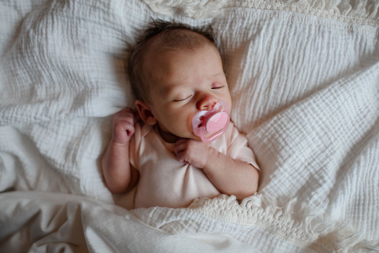 Sleeping newborn with pacifier lying on white textured bedding.