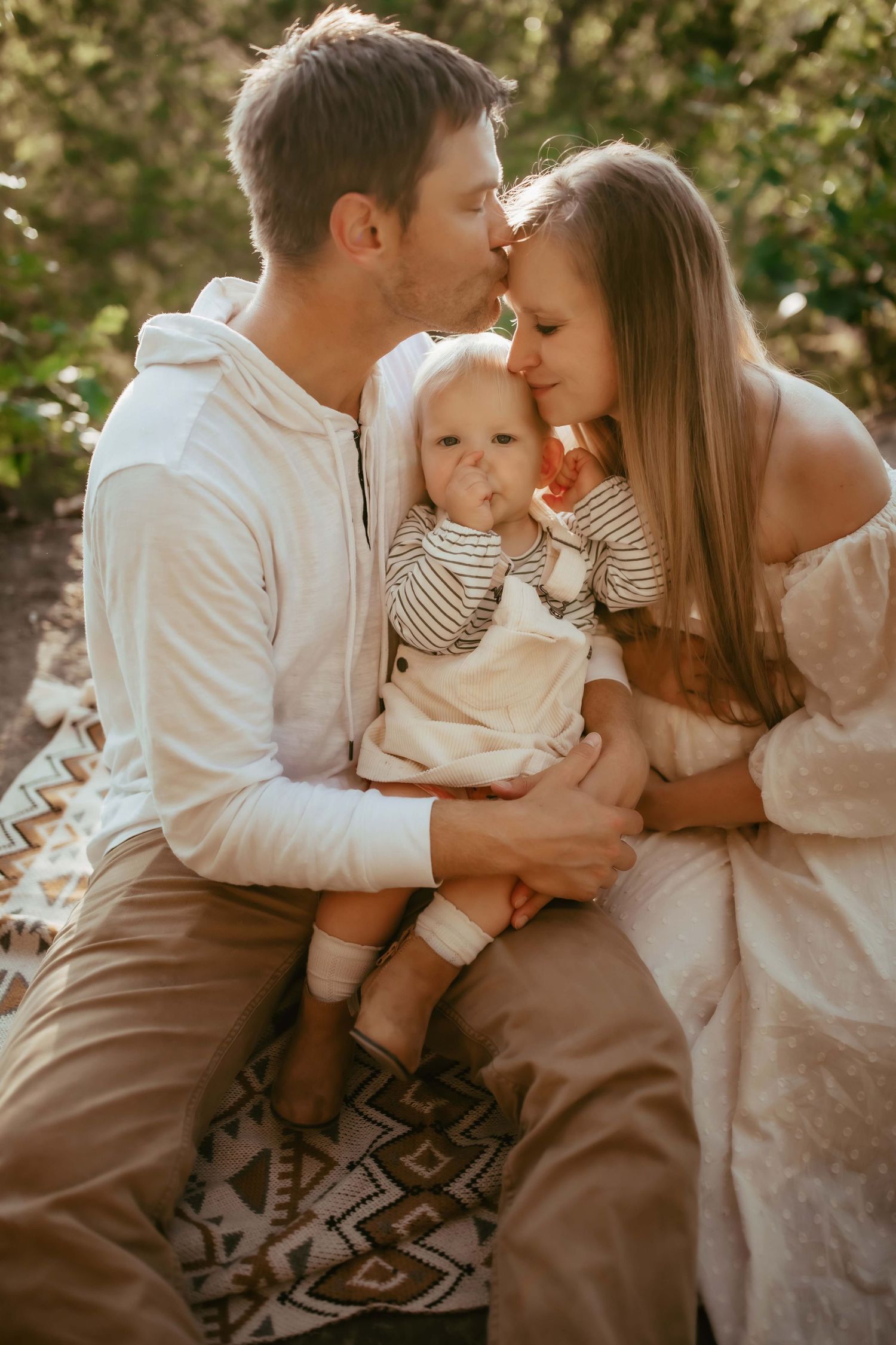 A loving family moment outdoors as parents embrace their small child while sitting on a geometric patterned blanket.