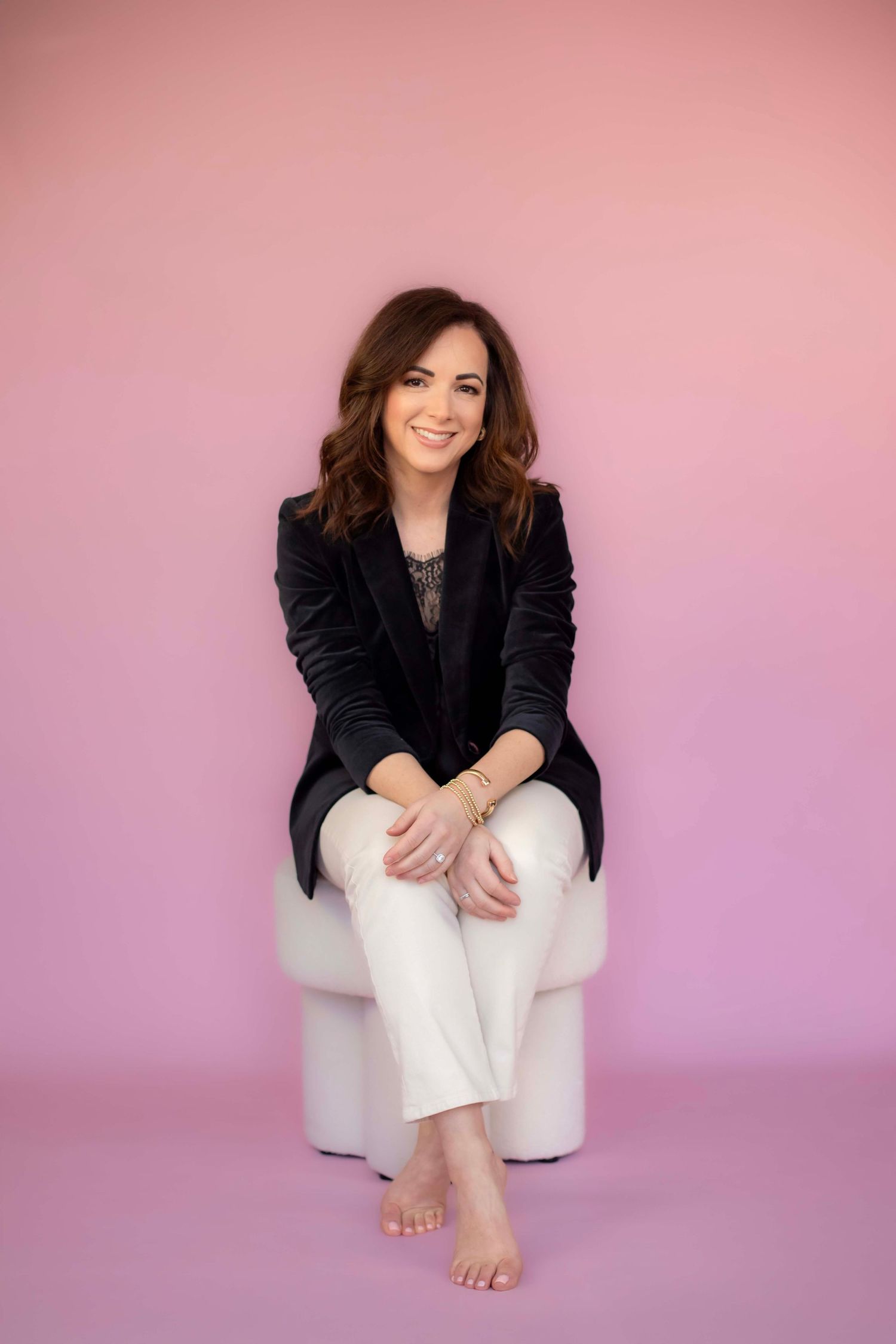 Casual professional seated portrait in black blazer and white pants against pink background.