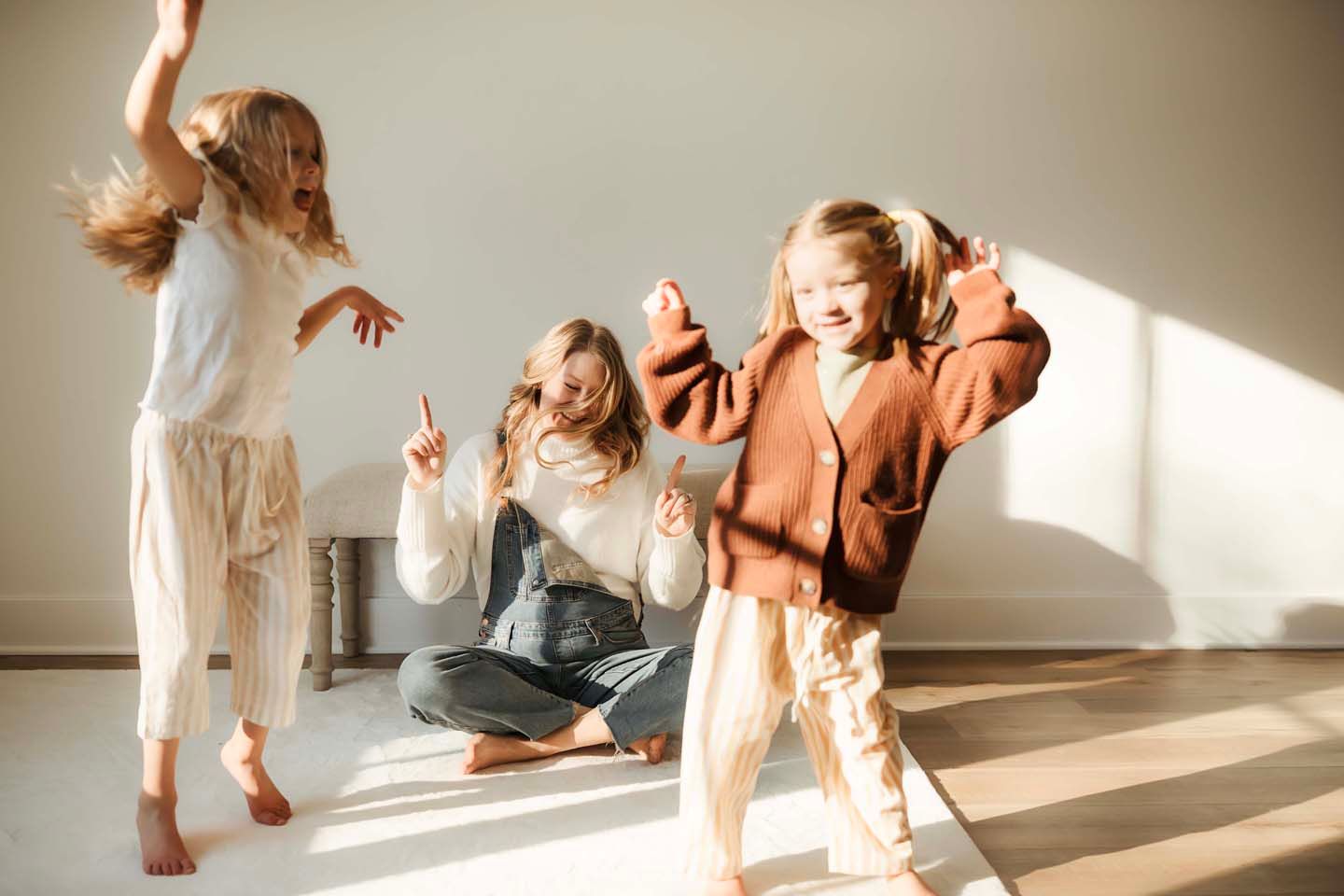 Three young girls dancing and playing joyfully in a sunlit room wearing neutral toned casual outfits.